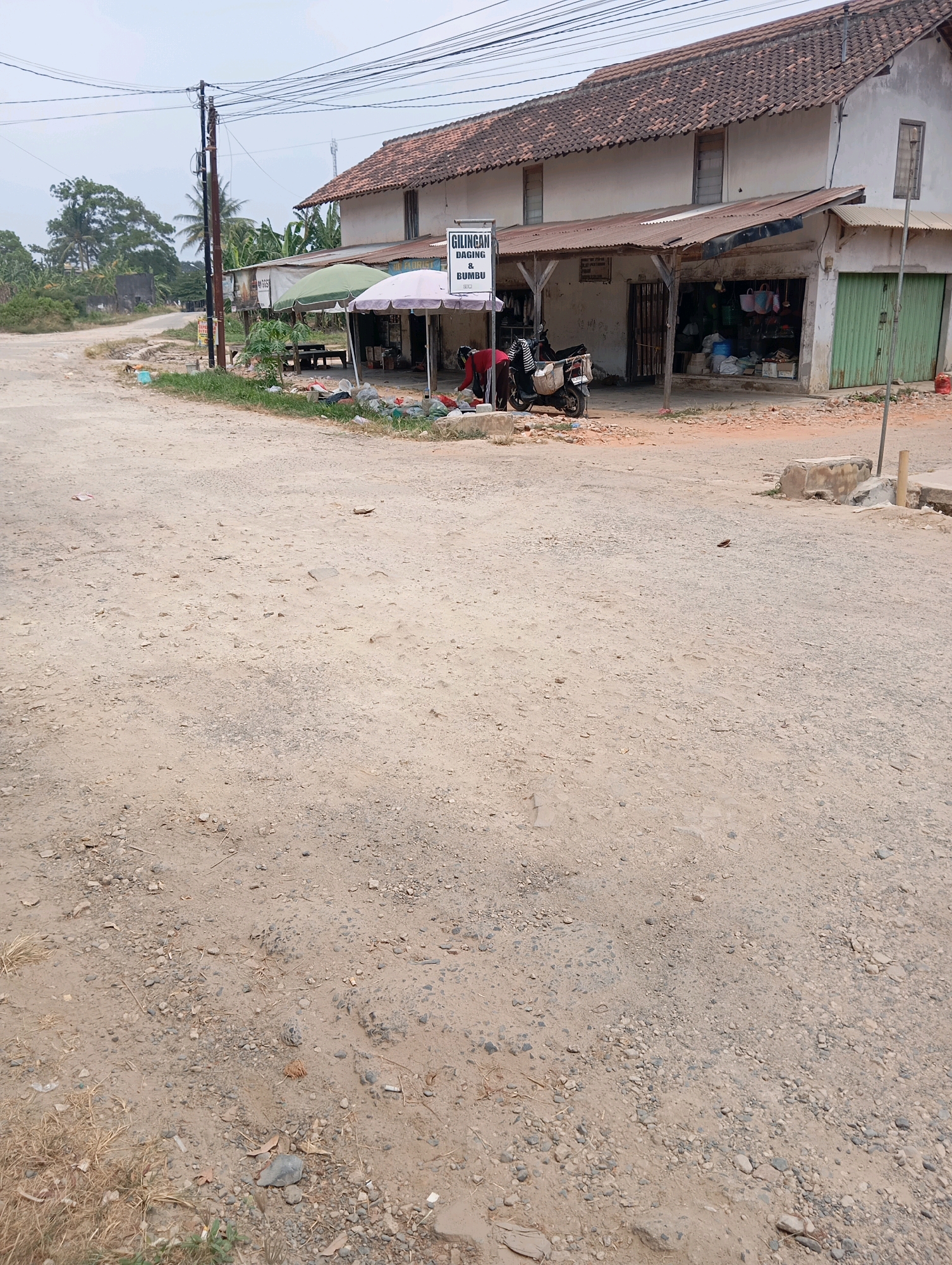 The image shows a street scene in Batang Hari Ogan, Indonesia. The street is unpaved and dusty, with gravel and dirt. On the right side of the street, there is a small building with a corrugated metal roof and a green metal garage door. Next to the building, there is a sign that reads "GILINGAN DAGING & BUMBU". In front of the building, there are several umbrellas providing shade over a market stall. A motorcycle is parked in front of the stall, and a person is partially visible, appearing to be engaged in some activity. The background shows more trees and a cloudy sky. There are also power lines overhead. The overall impression is of a rural or semi-urban commercial area.