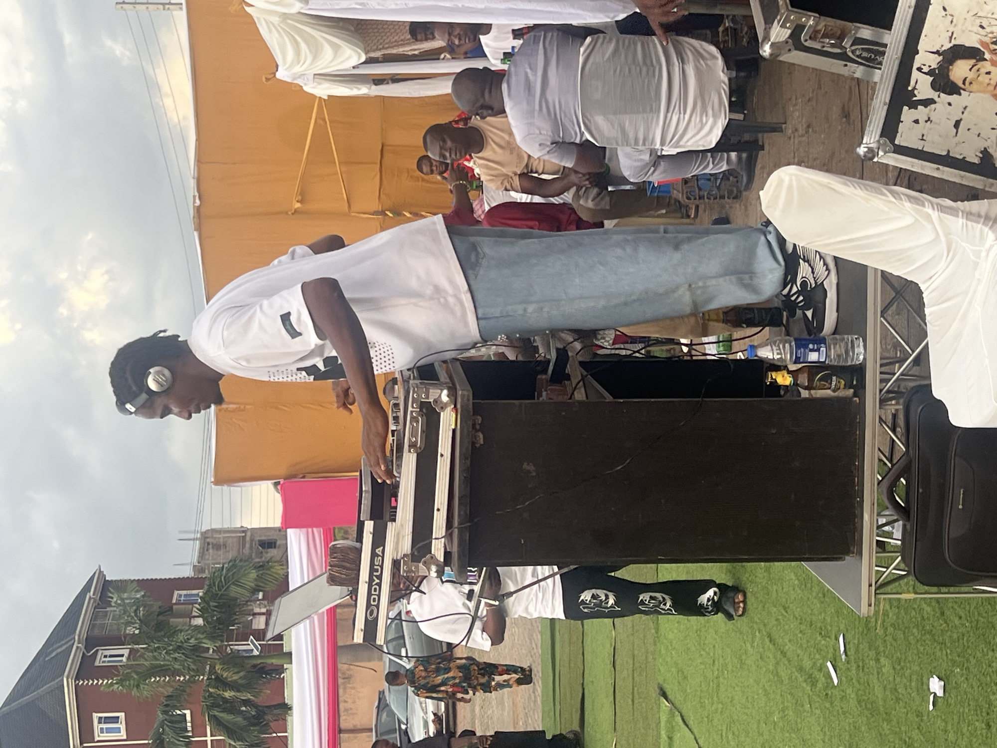 A medium shot captures a DJ at work in an outdoor setting. The DJ, wearing a white short-sleeved shirt, light blue jeans, and black sneakers, stands behind a black DJ console and wears headphones around his neck. He's focused on the equipment, suggesting active mixing or music selection.
Several other individuals are present in the background. Some are seated at tables under a tan-colored tent or awning, while others are standing. A building is visible in the background, and the presence of greenery indicates an outdoor venue.
There are cases and equipment near the DJ, one of which has a black-and-white portrait displayed on its front. A white chair is also present. The surroundings are somewhat cluttered with cables, water bottles, and other items associated with a DJ setup. The presence of draped materials in white and pink suggests the event might be celebratory, such as a wedding or a party. The location is Unknown, Unknown.