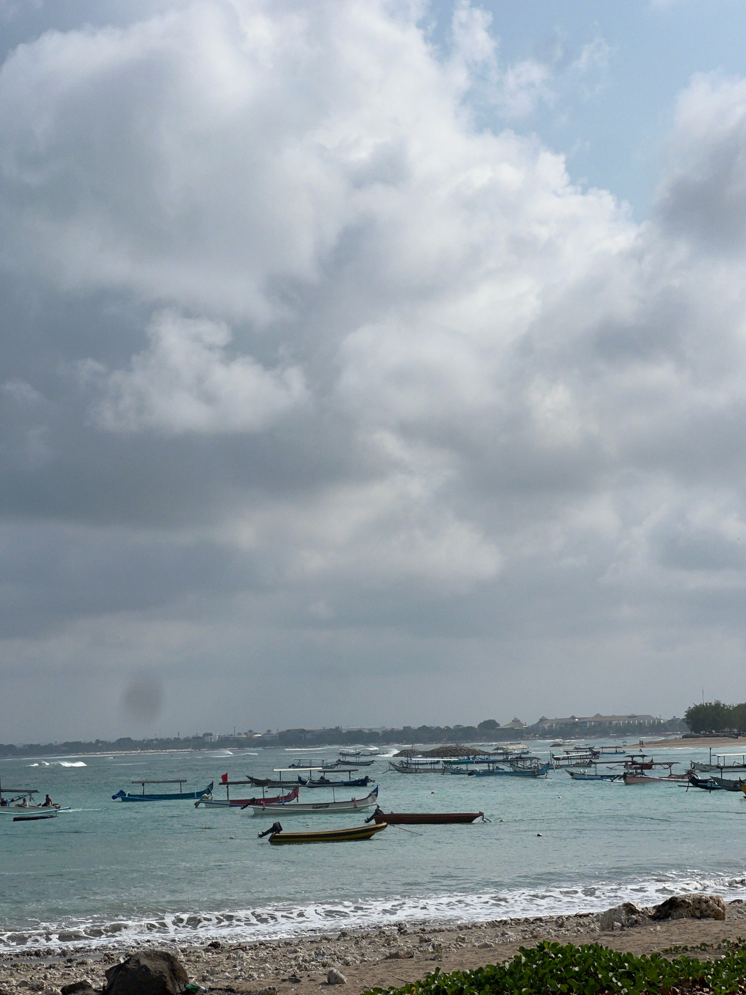 The image depicts a coastal scene in Kuta, Indonesia, characterized by a tranquil ocean dotted with numerous traditional Indonesian fishing boats. The water is a clear turquoise, with gentle waves lapping at the shore. The beach in the foreground is composed of small pebbles and rocks, with some green foliage visible.
In the middle ground, a flotilla of fishing boats, some with their characteristic outriggers, are anchored or drifting in the shallow waters. The boats are of various sizes and colors, with some appearing to be empty and others carrying a few people. The fishing boats are positioned in rows, suggesting a busy harbor or a waiting area for fishing activities. The boats' designs are consistent with those commonly found in Southeast Asian maritime communities.
The background reveals a distant shoreline with buildings and palm trees, hinting at the urban or resort development typical of Kuta. The sky is overcast with dramatic, heavy clouds, creating a subdued and somewhat moody atmosphere. The clouds occupy a significant portion of the frame, with patches of lighter sky visible. The overall impression is one of a serene, yet active, coastal environment in Kuta, Indonesia, where traditional fishing practices are still prevalent.