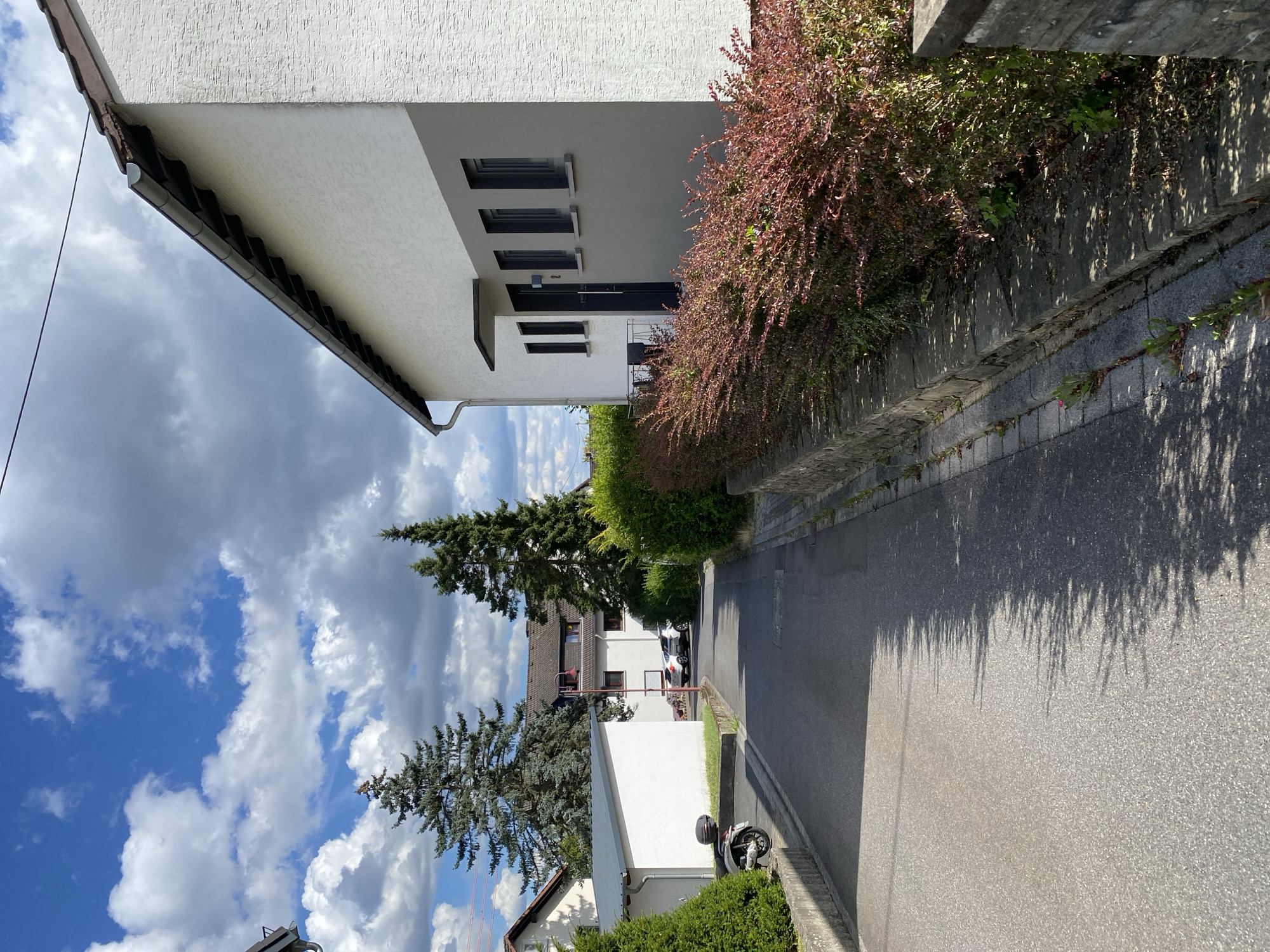 This is an outdoor, eye-level, medium-shot photograph of a street in the city of Flammersfeld, Germany, on a sunny day with scattered clouds. The street is paved and lined with residential buildings and greenery.
In the foreground, to the right, is the side of a white modern house with gray horizontal accents. It has a dark gray door and several dark-framed windows. A low stone wall runs along the front of the house, supporting a dense hedge with reddish-purple leaves.
To the left of the street, there is a white shed-like structure and a parked scooter. Further down the street, two cars are visible, a white SUV and a silver sedan. Tall evergreen trees stand behind the buildings, and the sky is blue with fluffy white and gray clouds. The street appears to be a residential area with a peaceful and tidy atmosphere.