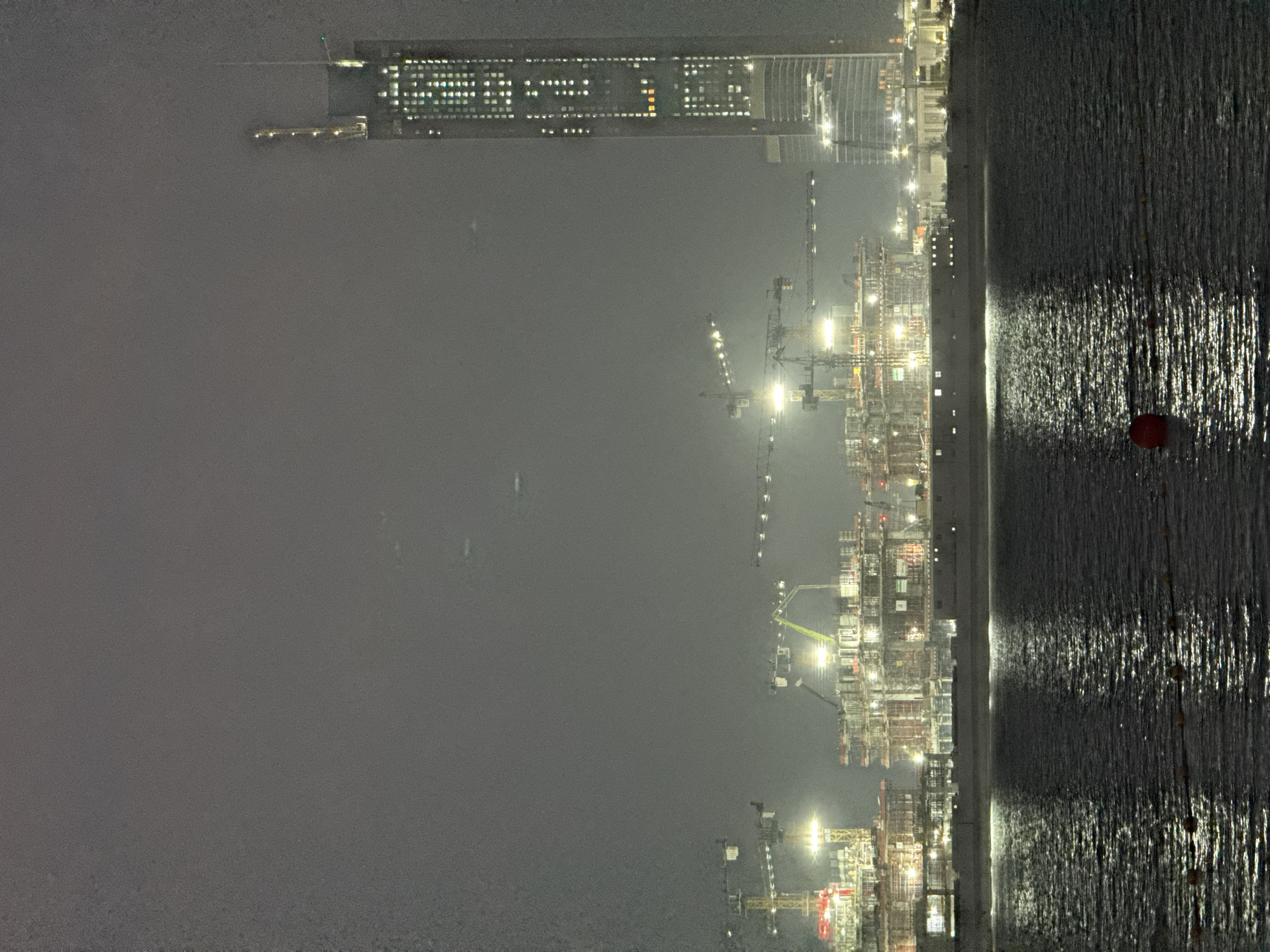 The image shows a nighttime view of a city skyline in Dubai, United Arab Emirates, dominated by a massive construction project. A very tall, slender skyscraper, illuminated by numerous windows, stands prominently on the right side of the frame. To the left, several large construction sites are visible, characterized by numerous cranes and partially built structures, all lit up. The construction areas appear to be extensive, with multiple levels and complex scaffolding. In the foreground, a body of dark water reflects the lights from the city and construction sites, creating shimmering patterns. A red buoy is visible floating on the water. The overall atmosphere is hazy, suggesting fog or a light atmospheric disturbance. No people are directly visible in the frame.