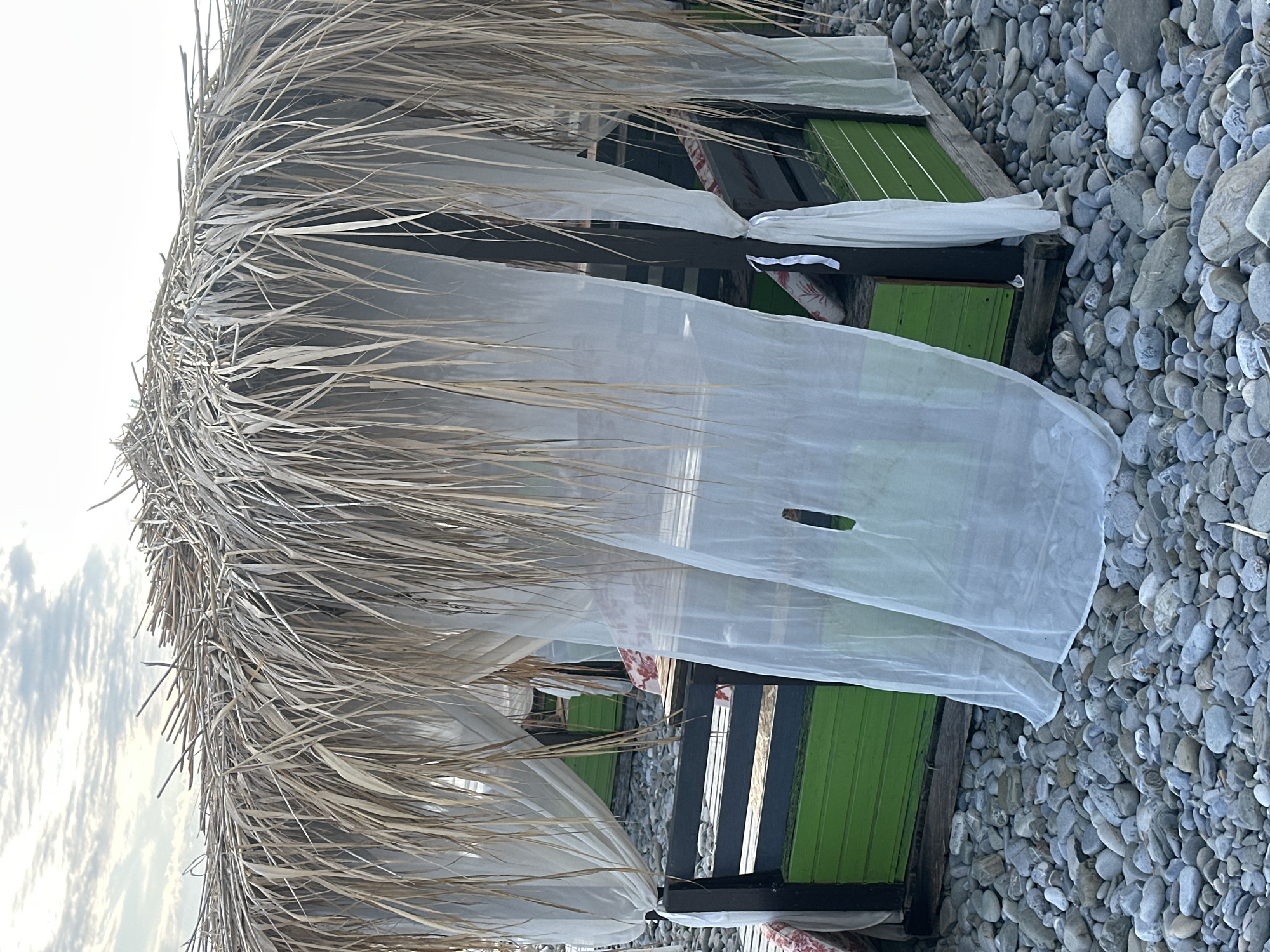 A beachside cabana with a thatched roof made of dried palm fronds is situated on a pebbly beach. White sheer curtains hang loosely from the cabana's wooden frame, partially obscuring the interior. The base of the cabana is painted bright green. The cabana is empty. The scene is set against a backdrop of a cloudy sky. This scene is located on the coast of Sochi, Russia.