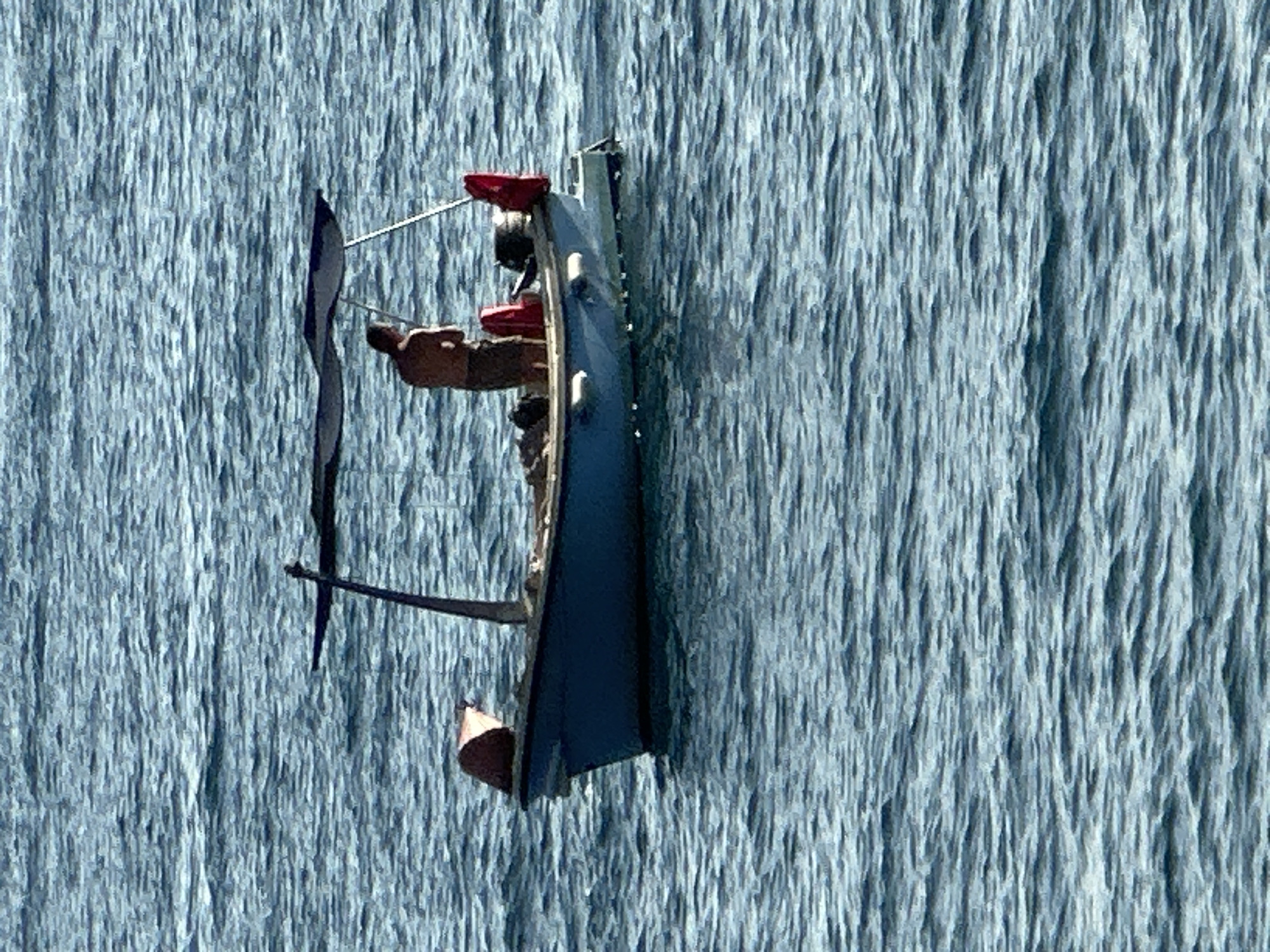 The media file shows a close-up, slightly tilted aerial view of a small boat with two people inside, navigating through a waterfall. The water is a vibrant blue, with vertical lines indicating the flow of the waterfall. The boat appears to be a dark blue hull with a brown interior and several red elements on top, possibly representing seating or accessories.
One person is visible in the boat, seated towards the front, wearing a dark top. Another person, possibly the rower, is partially visible further back. The activity taking place is navigating the boat down the waterfall. The water's texture suggests a fast-flowing current. The overall impression is one of adventure and a somewhat precarious journey. The lighting suggests it is daytime, with no harsh shadows. The backdrop of the waterfall is consistent with a natural setting, and the mention of Budva, Montenegro, suggests a coastal or riverine environment where such a waterfall might be found, possibly an attraction or a unique natural feature. No visible text is present in the image.