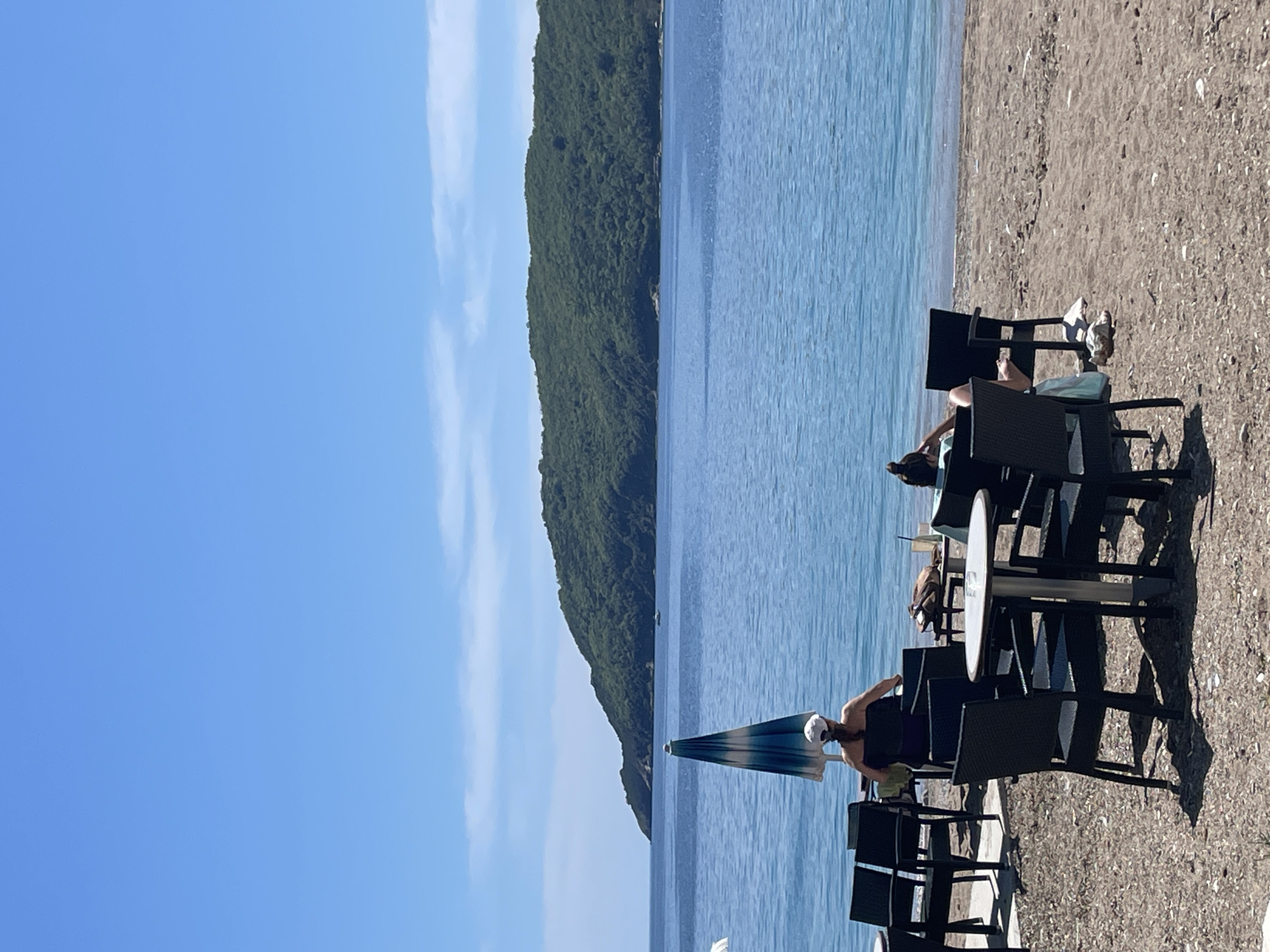 The scene captures a beach in Budva, Montenegro. The foreground features tables and chairs set up on a gravelly beach, with people seated at some of them. A partly-open blue and white striped beach umbrella stands near one of the tables. The middle ground consists of the Adriatic Sea, and across it lies a densely forested peninsula. A clear blue sky with a few wispy clouds stretches above.