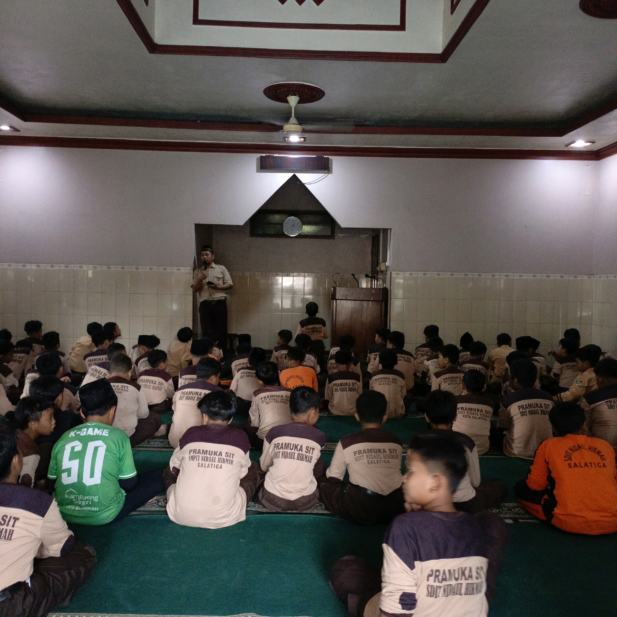 The image depicts an indoor gathering of approximately 50 children, appearing to be of elementary school age, seated on a green carpeted floor. A single adult male, dressed in a light brown uniform with a dark tie and cap, stands on the left side of the frame, holding a microphone and speaking to the children. In the background, a wall with light-colored tiled panels is visible, and a clock is mounted above a triangular architectural feature. The ceiling has a decorative design with red trim. Several children are wearing beige and white uniforms with "PRAMUKA SIT SDIT NIDAUL HIKMAH SALATIGA" printed on the back. One child in the front row is wearing a bright green t-shirt with "K-GAME 50" printed on it. The overall setting suggests an event or presentation taking place within a school or community building in Salatiga, Indonesia.