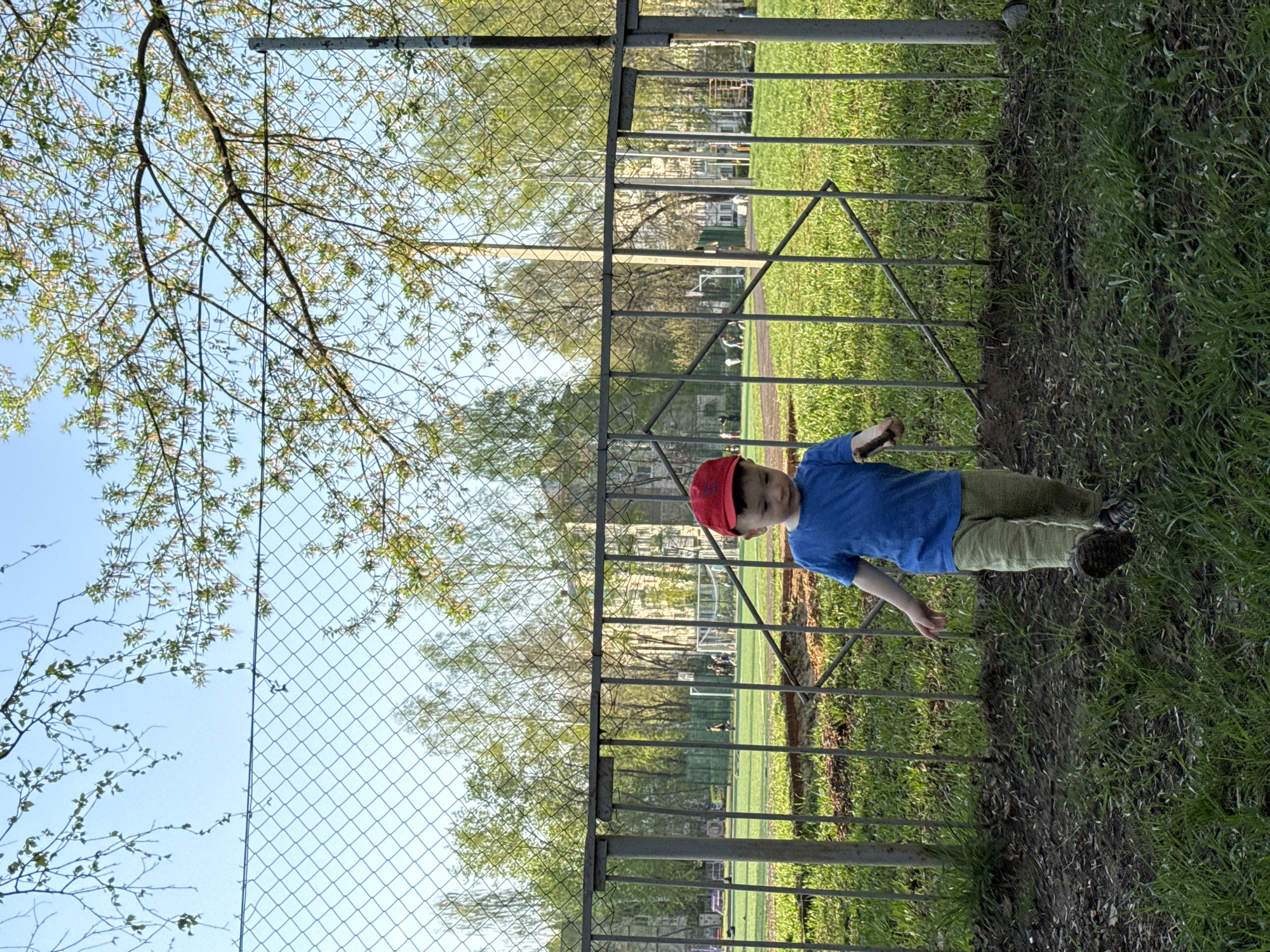 A young boy in a blue shirt, khaki pants, and a red cap stands near a metal fence bordering a grassy field. The field appears to be a sports field with soccer goals visible in the background. Beyond the field, buildings are discernible. The sky is blue with tree branches and leaves visible on the left side of the frame. A chain-link fence also runs alongside the metal fence. The location is likely Izhevsk, Russia, based on visual cues of the architecture and environment.