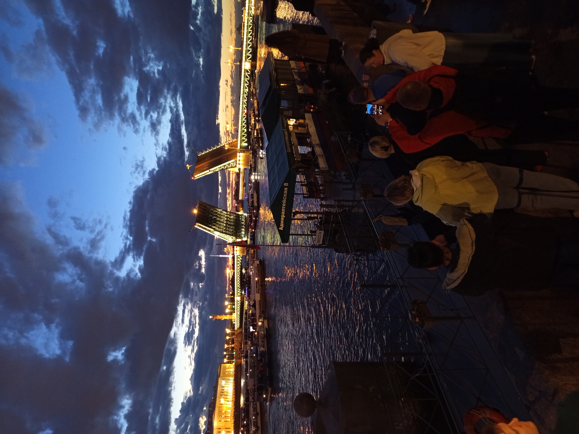 The image depicts a nighttime scene at a waterfront in an urban setting, possibly St. Petersburg given the context. A drawbridge, illuminated by lights, is raised in the background. The sky is dark with dramatic clouds, tinged with the orange glow of sunset or artificial lights. Several boats are visible on the water. In the foreground, a group of people are gathered on a promenade. Some are looking out at the scene, while one person is taking a photograph with a smartphone. The atmosphere suggests a tourist attraction or a popular viewing spot.