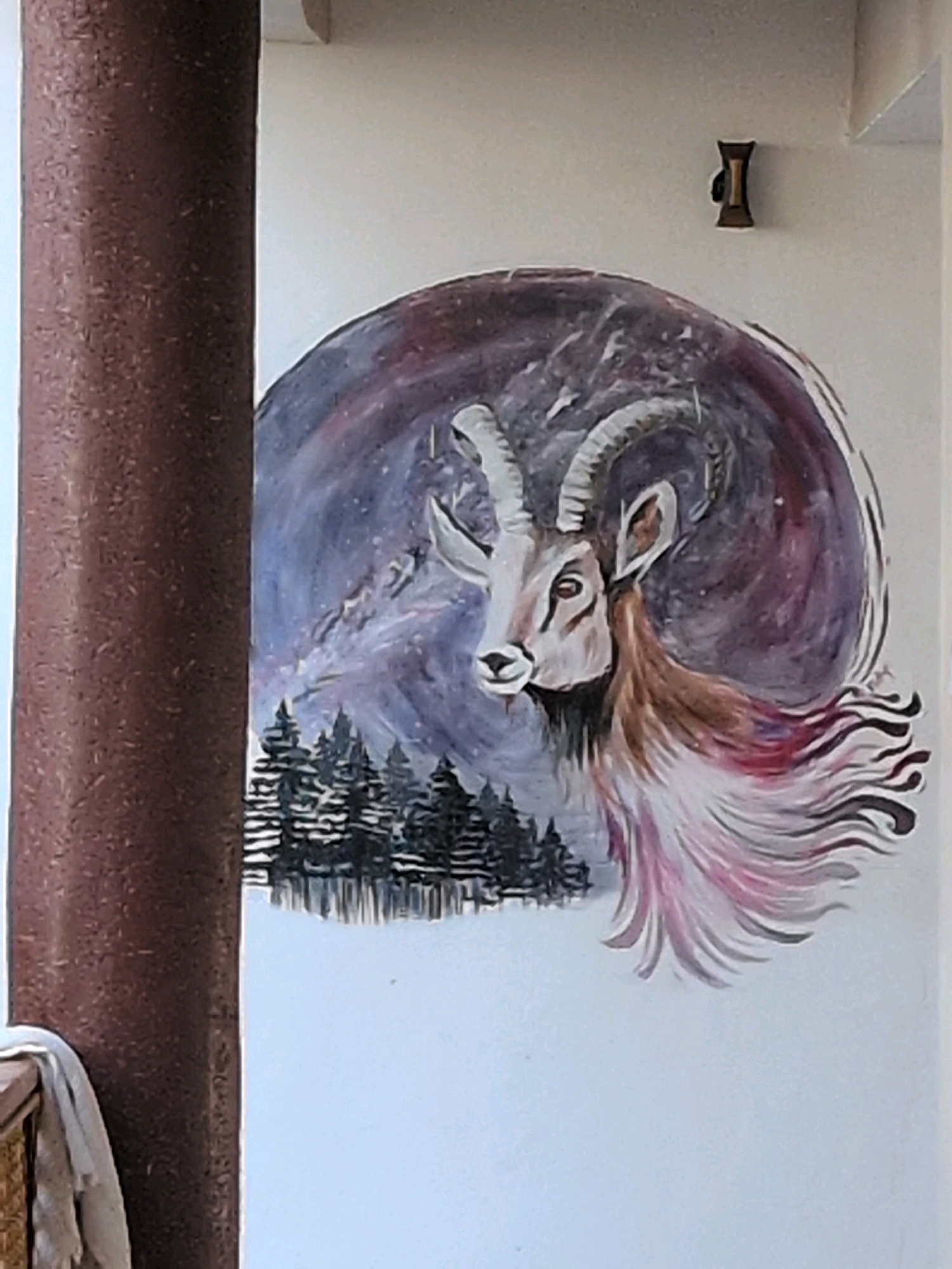 This image shows a mural painted on an exterior wall. The mural depicts a goat with large, curved horns, facing left. The goat's head is detailed, with an alert expression. Behind the goat, the mural transitions into a swirling, cosmic-like background of purples and blues, with hints of stars. In the lower portion of the mural, a dark silhouette of evergreen trees is visible, suggesting a mountainous landscape. The goat's mane is depicted as flowing, with streaks of pink and white, adding a dynamic element to the artwork. To the left of the mural, a large, cylindrical, dark brown pillar is present, partially obscuring the left side of the artwork. A white cloth is draped over a railing in the lower left corner of the image. A small, dark, hourglass-shaped object is mounted on the wall above and to the right of the mural. The image appears to have been taken in the city of Leh, India, judging by the architectural style and the presence of what seems to be a traditional Ladakhi element in the mural's design, likely a decorative feature.