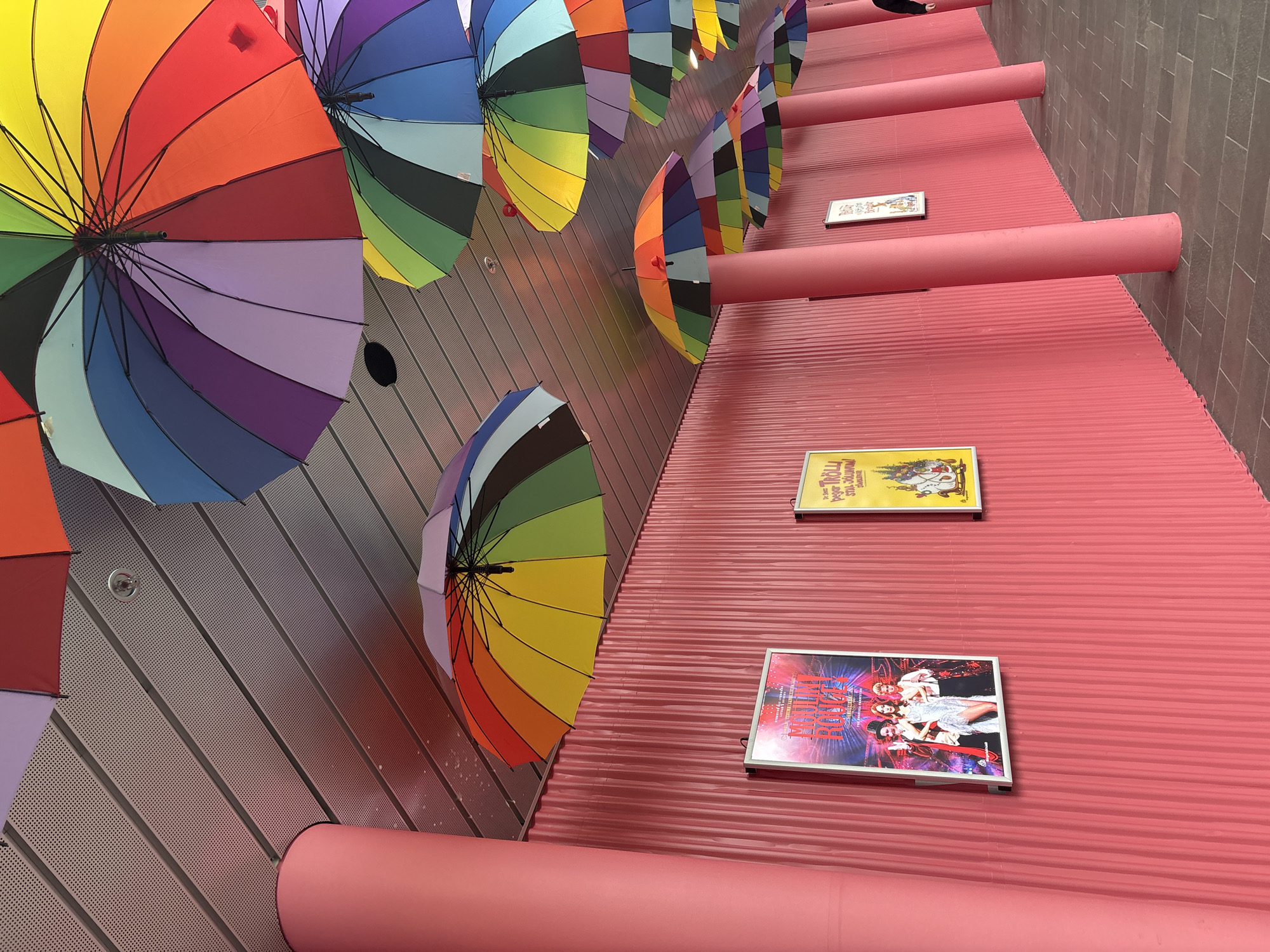 The image depicts a covered walkway in Reykjavik, Iceland, characterized by a vibrant and whimsical aesthetic. The walkway features a corrugated pink wall on the right and a series of pink cylindrical pillars supporting a metallic ceiling. Hanging from the ceiling, a multitude of colorful umbrellas, arranged in a rainbow pattern, create a canopy overhead.
On the pink wall, several framed posters are displayed. The most prominent, on the left, is a brightly colored poster advertising "Moulin Rouge!" with images of three performers. To its right, a yellow poster features cartoon characters. Further down the wall, additional posters are visible, though less distinct.
The ground of the walkway is paved with dark grey interlocking tiles. A person, partially visible on the far right, is walking away from the camera, dressed in dark clothing. The overall scene suggests a public space decorated for a festive or artistic event, with the colorful umbrellas providing a striking visual element against the pink backdrop.
