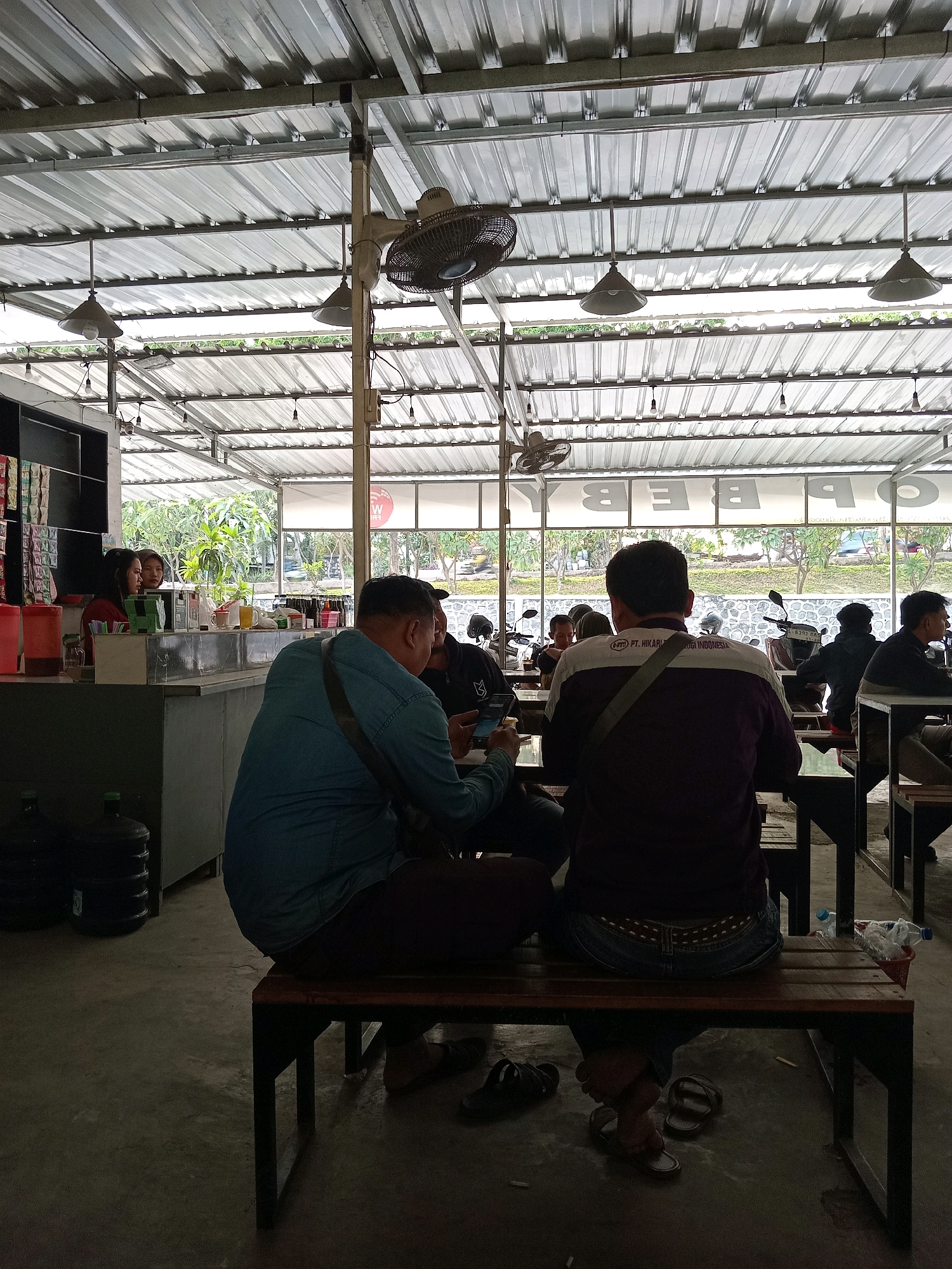 The image depicts an outdoor seating area, likely a food stall or cafe, with a corrugated metal roof supported by metal beams. Several people are seated at wooden tables and benches. In the foreground, two men are facing away from the camera, seated on a bench. One man wears a denim shirt, and the other wears a purple and white jacket with a logo that reads "PT. HIKARI INDONESIA". To the left, a counter area is visible with two women standing behind it, and bottles of drinks on display. In the background, a line of motorcycles is parked, and beyond that, lush green trees and foliage are visible, suggesting an outdoor, possibly rural or semi-rural, setting. The overall atmosphere appears casual and relaxed, with a combination of natural light filtering through the roof and artificial lighting. The location is identified as Randuagung, Indonesia.