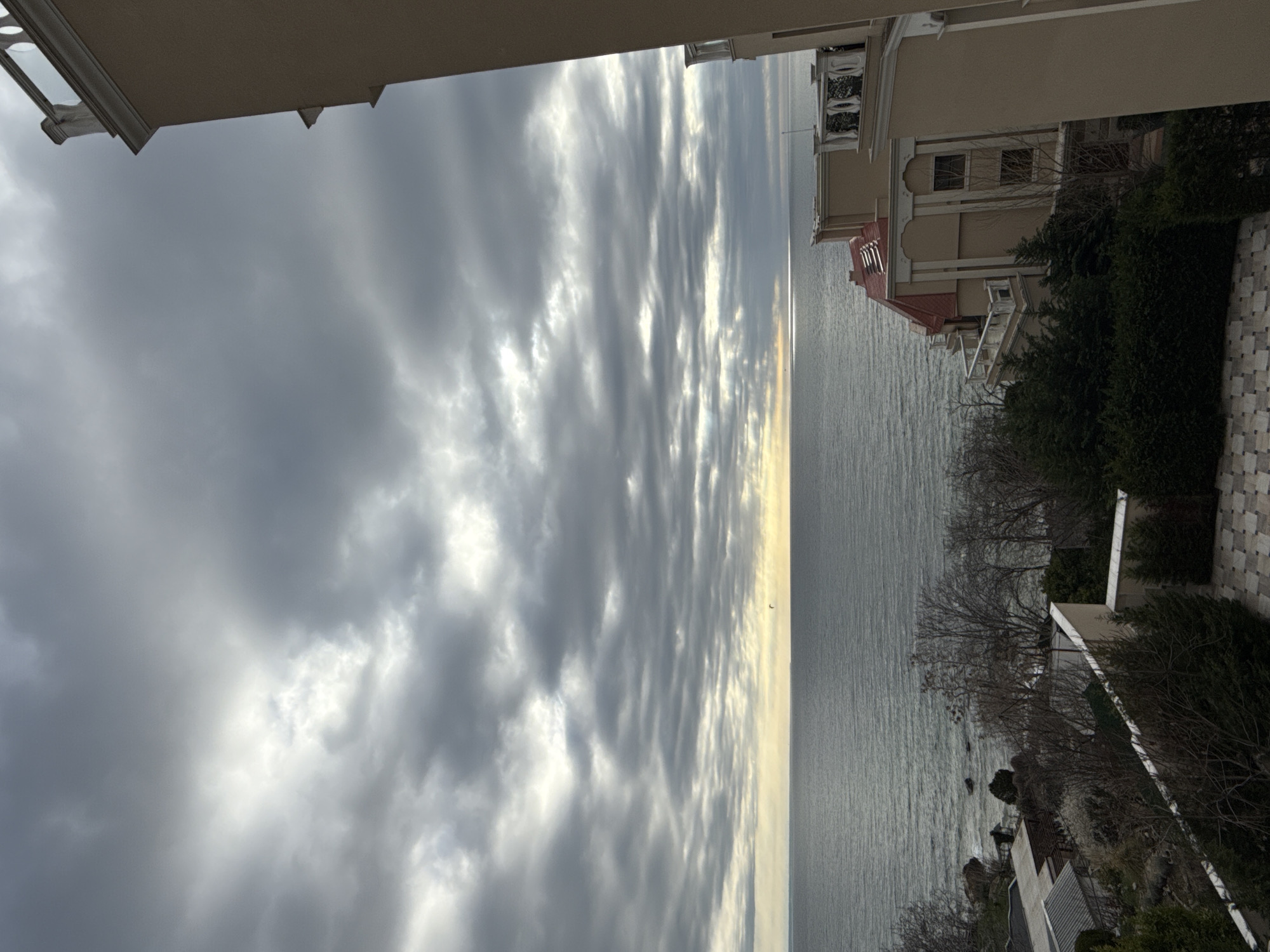 The image captures a view of the Black Sea from what appears to be a high-rise building or cliffside property in Varna, Bulgaria. The sea is visible in the center of the frame, with the coastline extending along the right side. The sky is overcast with dramatic, textured clouds covering much of the upper portion of the image. Buildings with distinctive architecture are seen to the right, partially obscured by trees and vegetation. A stone-tiled terrace or walkway is also visible. The overall scene suggests a residential or resort area near the coast on a cloudy day.