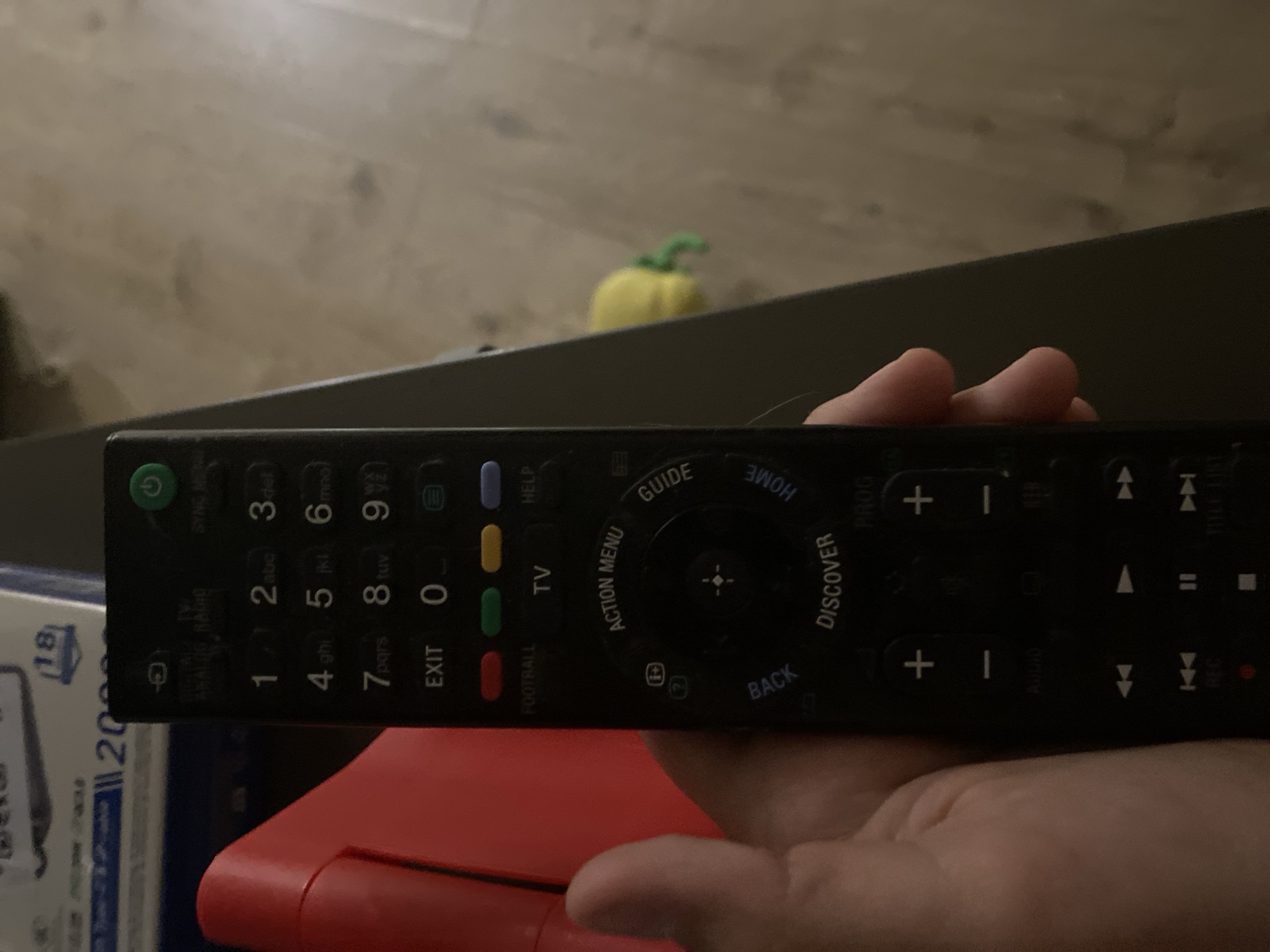 A person's hand is holding a black remote control. The remote control has various buttons, including numbers, power, and navigation controls. The background appears to be an indoor setting with some objects visible, including a red object to the left and a yellow object in the background. The image does not contain enough information to determine the location in Tolyatti, Russia.
