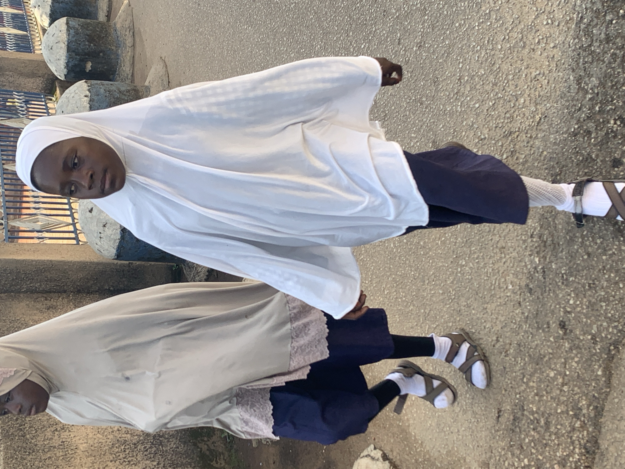 Two young girls are walking on a paved street, likely in Jos, Nigeria. The girl in the foreground is wearing a white hijab, a navy blue skirt, white fishnet stockings, and brown sandals. She is looking to her left. The girl behind her is wearing a light beige hijab with a pink lace trim, a navy blue skirt, black leggings, and brown sandals. She is also looking to her left. They are walking in the same direction. The background shows a textured wall, a metal fence, and decorative concrete bollards. The ground is a rough, asphalt-like texture. The lighting suggests it is daytime, possibly late afternoon.