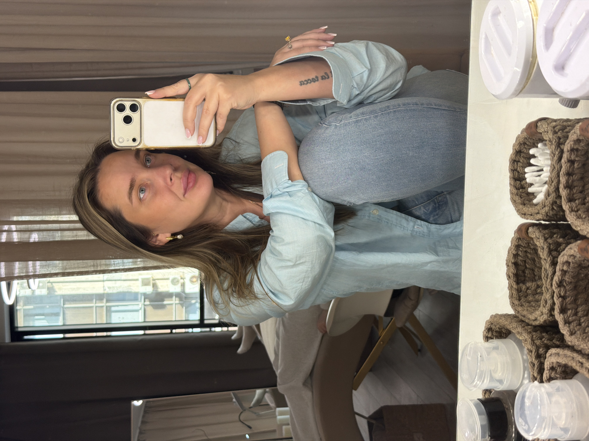 A woman is taking a selfie in a mirror. She is wearing a light blue button-down shirt and blue jeans. She is sitting on a chair, with her legs crossed. Her hair is blonde and she is wearing a ring on her finger. In the background, there is a window with a view of buildings in Moscow, Russia. On a shelf on the right side of the mirror are what appear to be baby products.