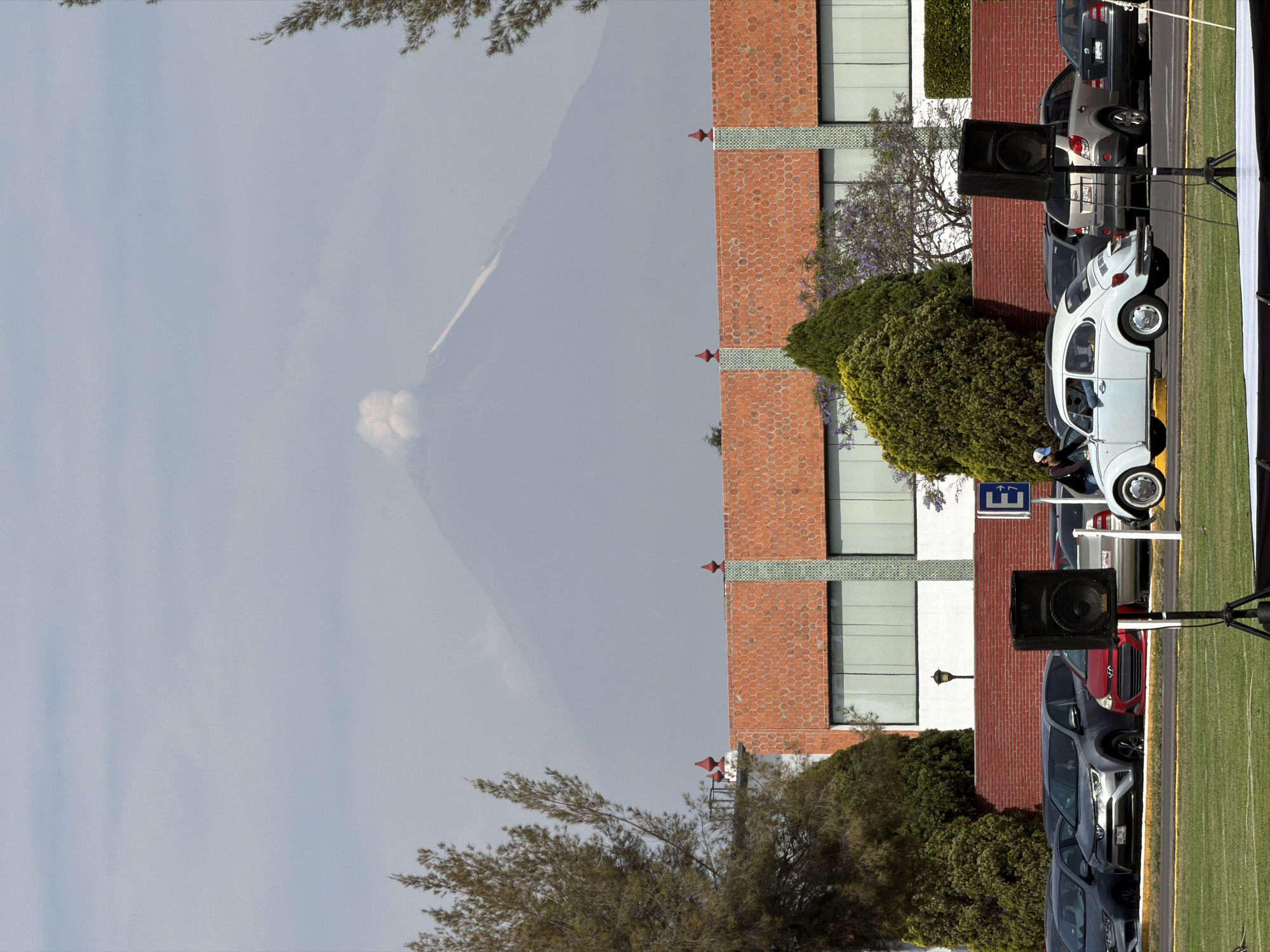 The image captures a scene in San Andrés Cholula, Mexico. In the background, a dormant volcano looms under a grey sky. Closer to the foreground, there's a building constructed with red brick. To the right side of the image, a line of cars are parked parallel to the building, with a white vintage car being especially noticeable. Several speakers stand near the cars, presumably for some event. A green field is at the bottom right.