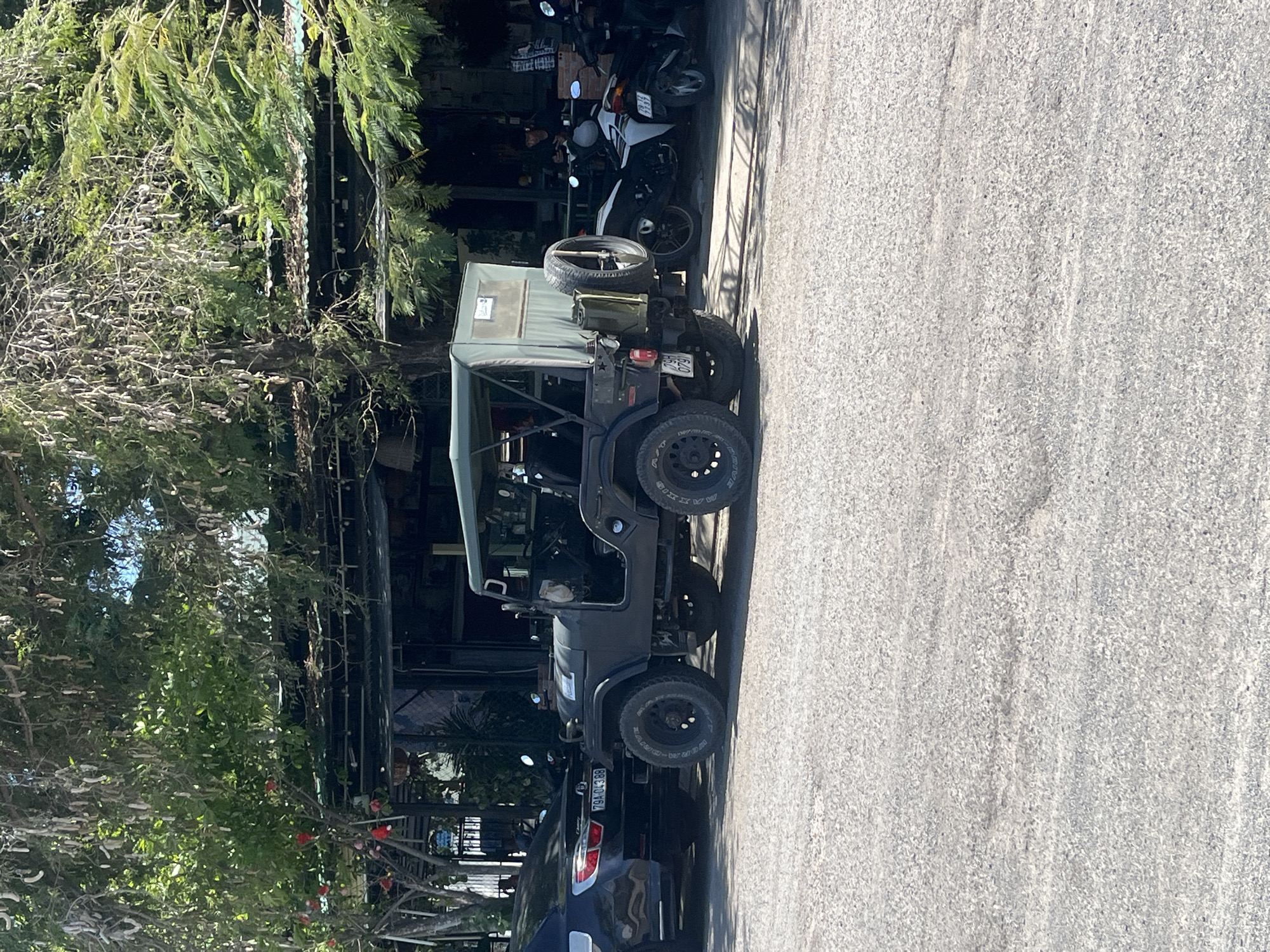 The scene captured is a street view in Bac Nha Trang Ward, Vietnam. A black Jeep with a spare tire mounted on the back is parked on the side of the road. The vehicle has a dark blue or black exterior and rugged tires. Part of a blue car is parked directly behind the Jeep. To the left of the Jeep, there's a commercial establishment, possibly a cafe or restaurant, partially obscured by dense foliage including flowering trees with red blooms. Several motorcycles are parked in front of the commercial space, further adding to the urban street setting. The road is paved and appears to be relatively clean and well-maintained. The angle of the image makes the scene appear on its side.