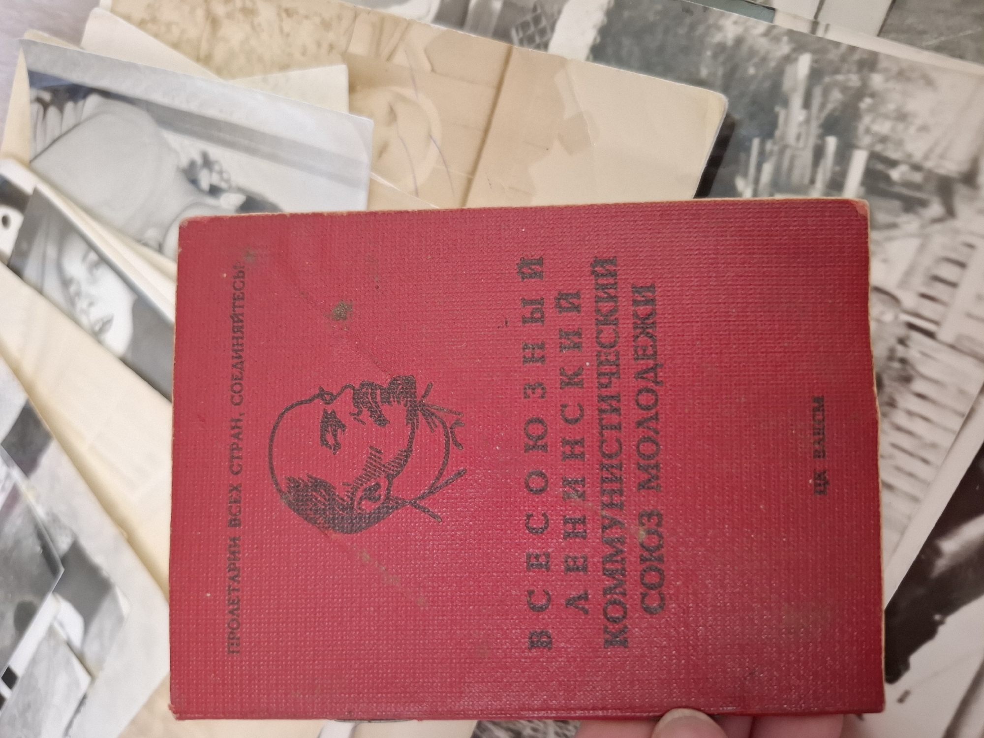 The image shows a red book on top of a pile of photographs. The book has the emblem of the Komsomol (Young Communist League) and text in Russian, indicating it's a Komsomol membership card. The phrase "Proletarians of all countries, unite!" is also visible. The book mentions "Комсомол Молодежи", which translates to "Komsomol Youth". The text "ГК ВЛКСМ" is also present on the book. There is no explicit mention of the location "Амзя, Russia" in the visible part of the book.