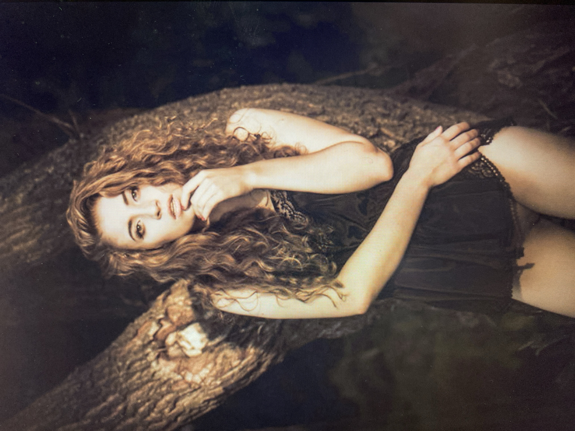 A woman with long, curly brown hair stands with her back to a tree trunk in what appears to be a dimly lit, outdoor setting. She is wearing a black, lacy slip dress that reveals her legs. Her right hand is raised to her chin, and her left arm rests on her hip. The lighting suggests it is nighttime or a very shaded area. The background is dark and out of focus, with hints of foliage.