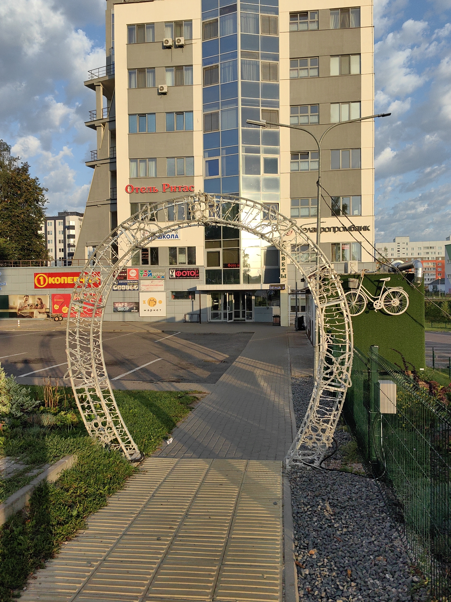 In Fanipal, Belarus, a modern multi-story building with cream and gray facades stands tall, featuring numerous windows and balconies. In front of the building, a large, white, illuminated archway structure is prominently displayed. The archway is adorned with a multitude of small lights, creating a decorative effect. To the right of the archway, a green, artificial turf wall is decorated with a white bicycle sculpture. The ground is paved with interlocking bricks, and a walkway leads towards the building's entrance. A metal fence encloses the area to the right, with landscaping visible beyond it. The sky above is a mix of blue with scattered white clouds, illuminated by natural sunlight, casting long shadows on the paved areas. Several signs are visible on the building's facade, indicating businesses and services within, including "Отель Ригас" (Hotel Rigas) and "Белагропромбанк" (Belagroprombank). A streetlamp is also present near the building.