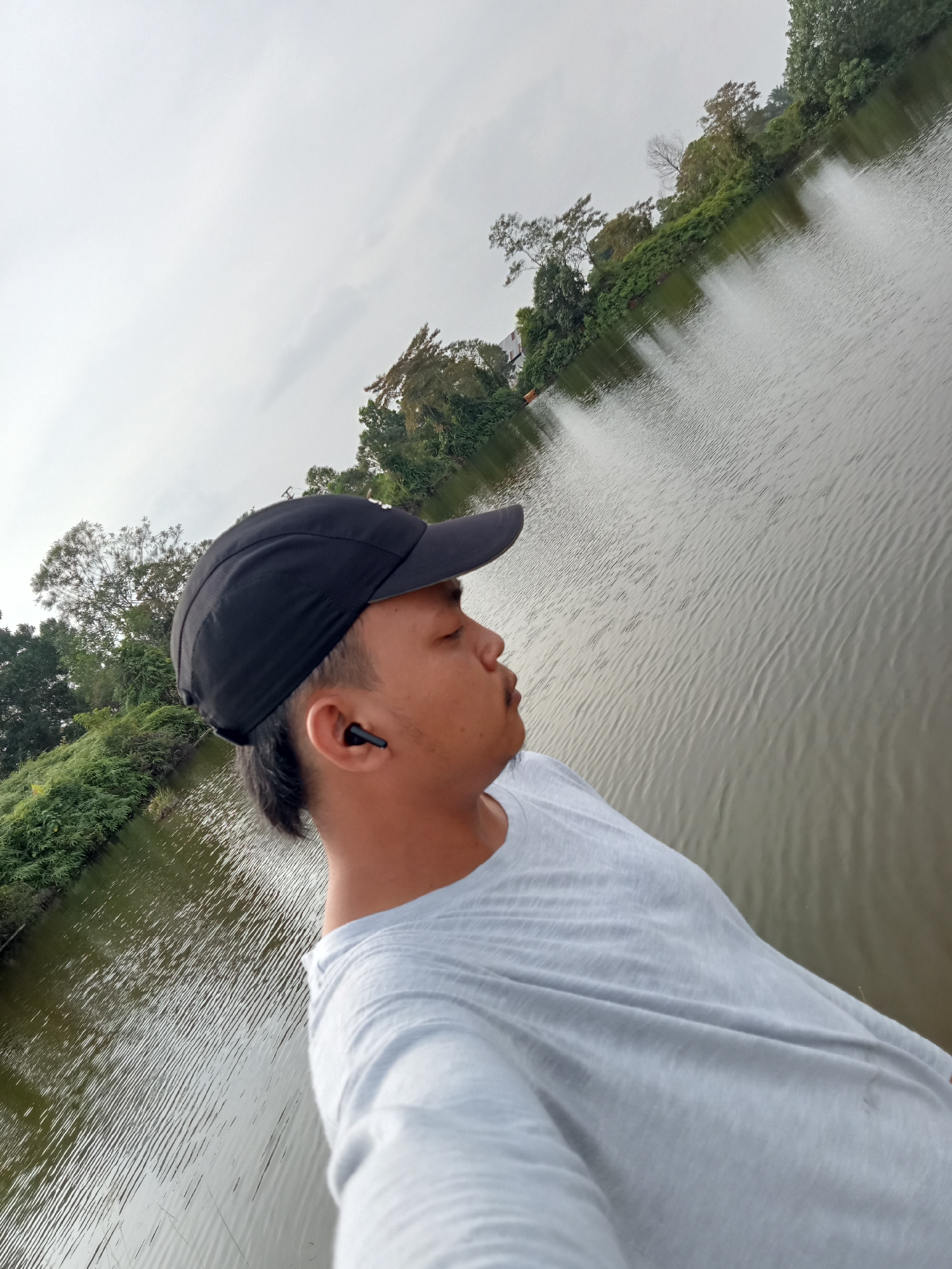 The image shows a person wearing a black baseball cap and a light gray t-shirt, looking to the right with their eyes closed. They have an earbud in their right ear. The person is standing on a platform next to a body of water, which appears to be a lake or a river. The water has ripples on its surface and reflects the overcast sky and the surrounding greenery. Lush vegetation, including trees and bushes, lines the edge of the water. The background shows more trees and foliage, with a cloudy sky overhead. The location is identified as Kualu, Indonesia, suggesting an outdoor setting within or near the city. The scene depicts a moment of quiet contemplation or relaxation by the water.