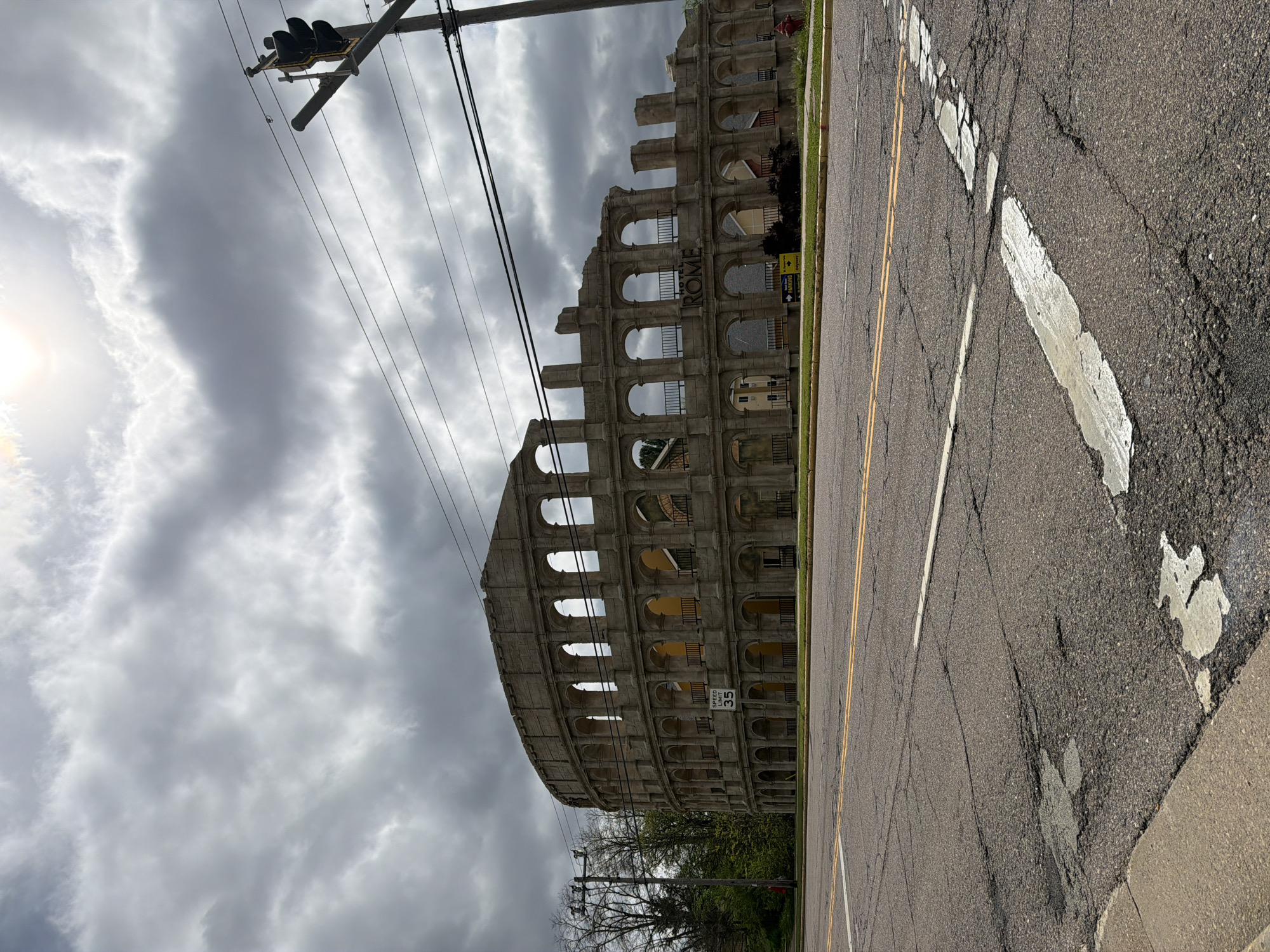 The image shows a roadside view of a partial replica of the Roman Colosseum, located in Wisconsin Dells, Wisconsin, USA. The structure is situated adjacent to a street with yellow and white painted lines. Overhead power lines and a traffic light are visible. The sky is overcast with heavy cloud cover. The facade of the Colosseum replica has multiple archways and architectural detailing. There are parking signs posted next to the structure. A speed limit sign is also visible.