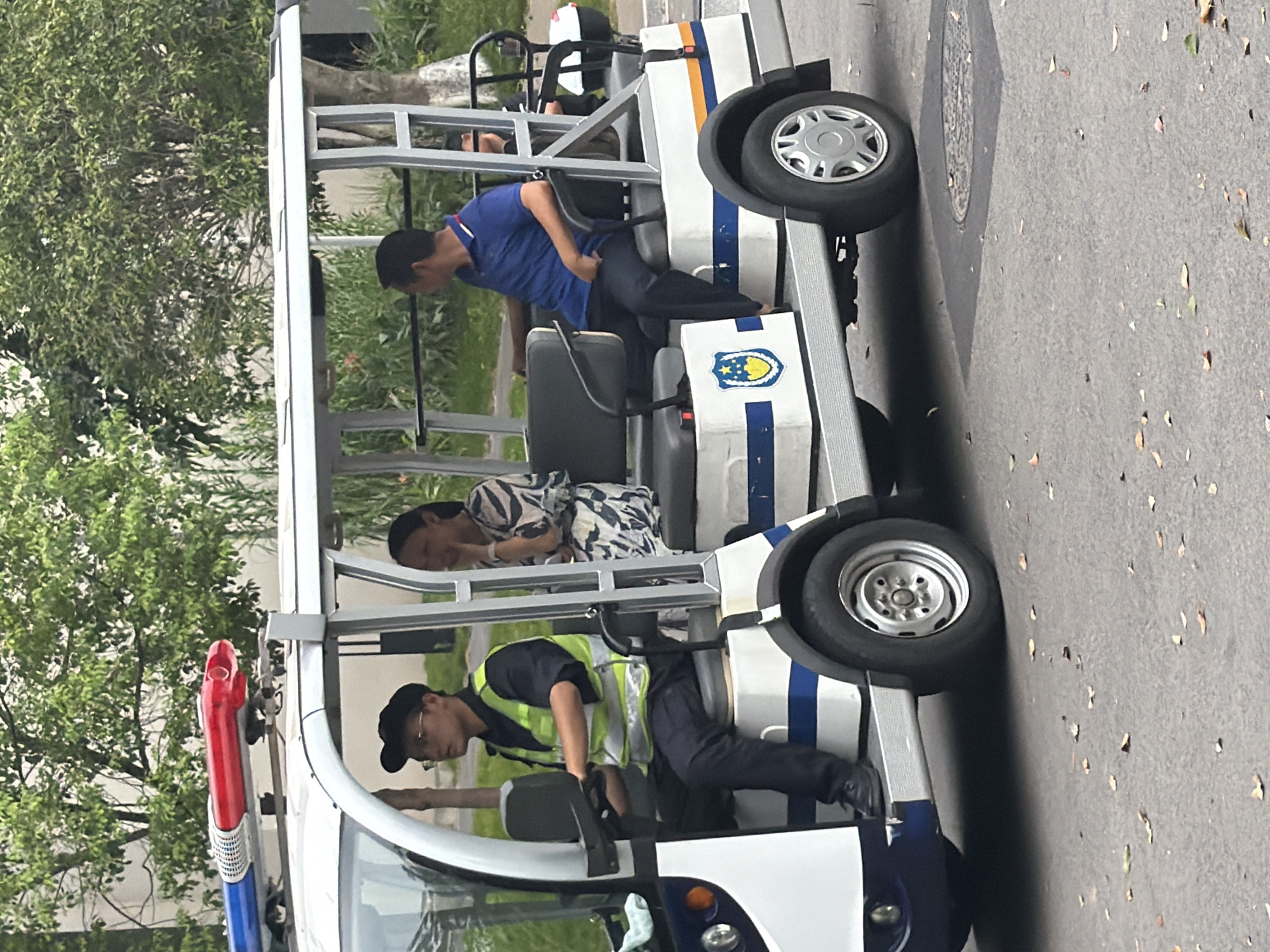In Jiangjin, China, a white electric utility vehicle with a light bar containing red and blue lights is parked on a paved road. The vehicle has a canopy and two rows of seats. The driver, a young man wearing a black cap, glasses, and a neon yellow vest over a dark shirt and pants, is seated in the driver's seat. To his right, in the second row, a woman wearing a patterned dress is seated, with her hand near her face. Further back, a man in a blue polo shirt and dark pants is seated. The vehicle is decorated with blue and yellow stripes. A blue and gold shield emblem is visible on the side of the vehicle. Trees and foliage are present in the background. The road surface is asphalt with scattered leaves.