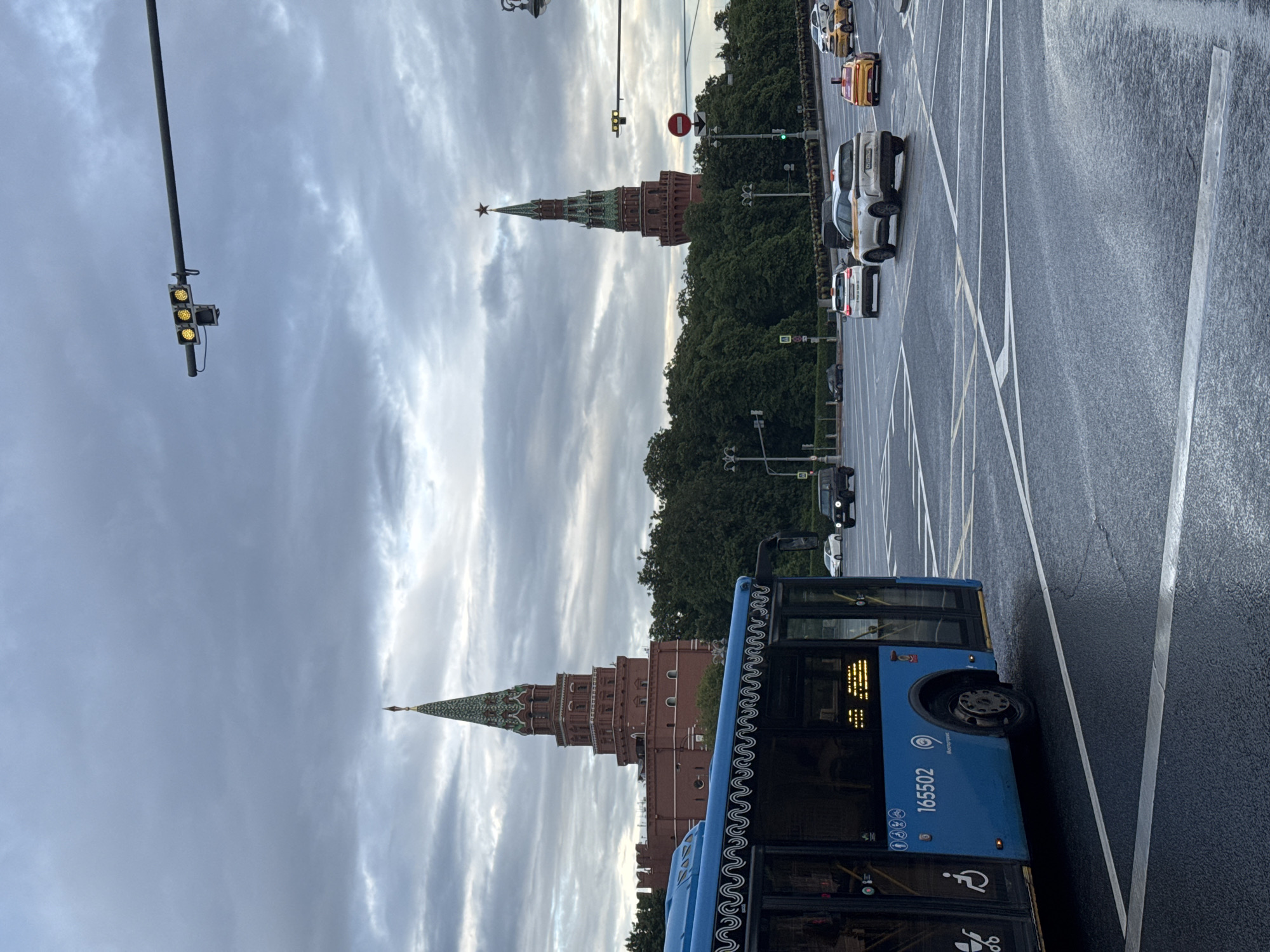 The image depicts a street scene in Moscow, Russia, featuring a blue city bus with license plate number 165502 in the foreground. Behind the bus, the towers of the Moscow Kremlin are visible, set against a cloudy sky. A traffic light on a pole stands to the left of the Kremlin towers. The street has visible white lane markings, and there are several cars and taxis traveling along the road in the background. Trees line the street, partially obscuring some of the Kremlin towers.