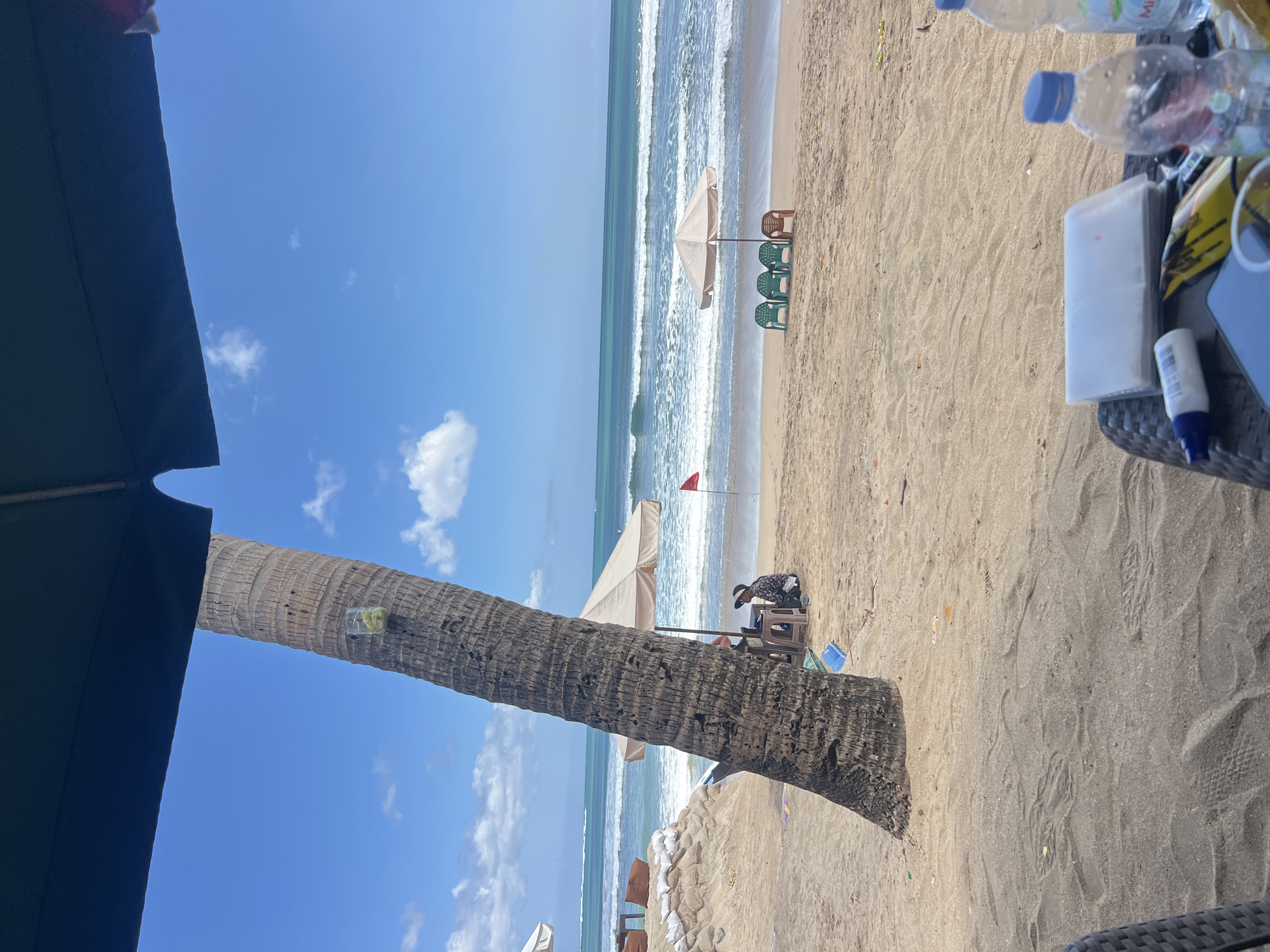 The image depicts a sunny day at Kuta Beach, Indonesia, with the ocean in the background. A large palm tree stands prominently in the foreground, leaning towards the left. Under a tan umbrella, a person wearing a hat and patterned clothing sits on a small stool next to a table. In the mid-ground, three green chairs and one brown chair are arranged under another tan umbrella. A red flag is planted in the sand behind the chairs. The beach is sandy, with footprints visible in the foreground. On a table in the foreground, several items are present, including two plastic water bottles, a white tube, a stack of papers, and a smartphone. The sky is bright blue with scattered white clouds.