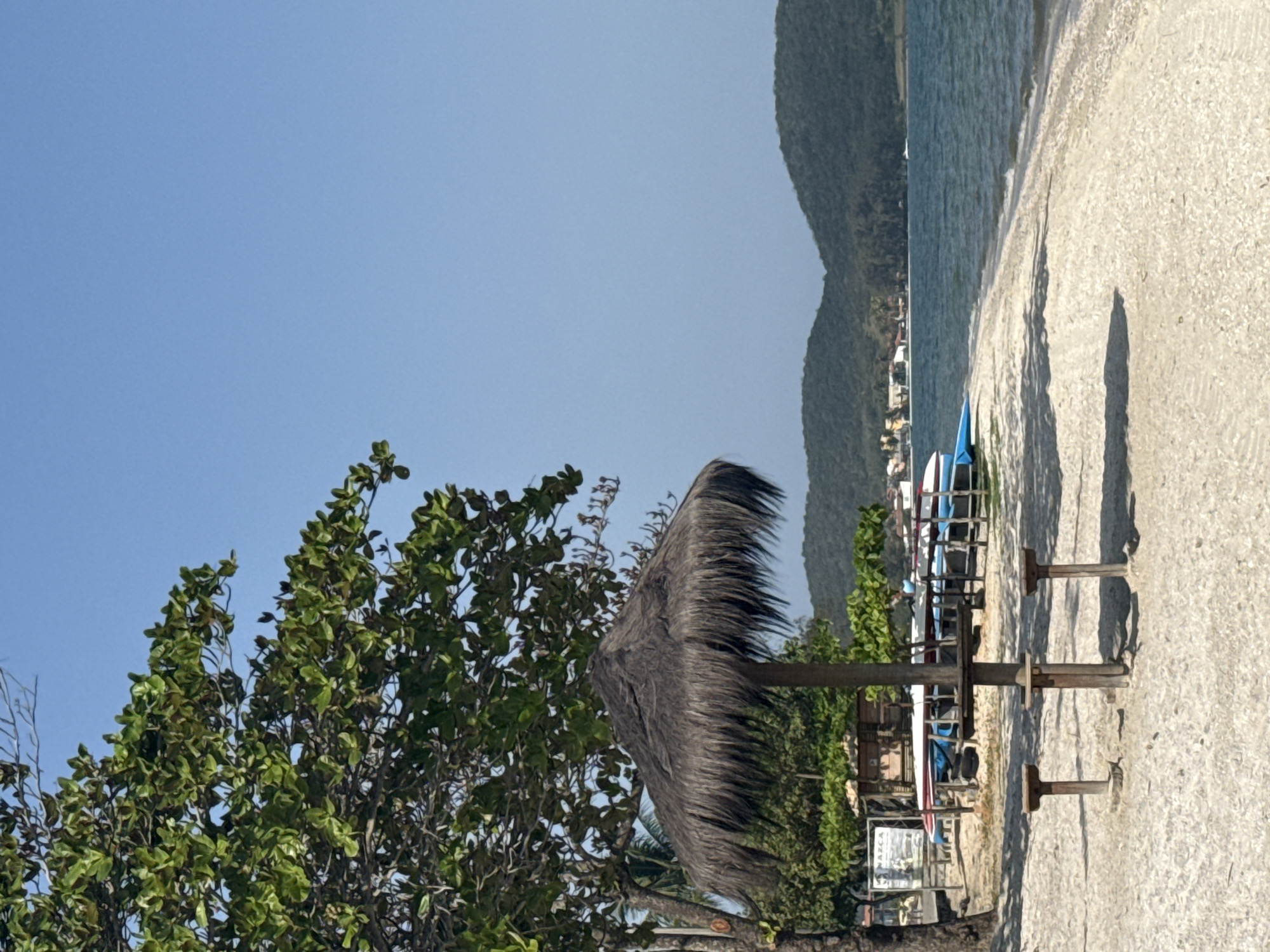 This is a medium-shot, eye-level photograph of a tropical beach scene in Iguaba Grande, Brazil, under a clear blue sky. The foreground is dominated by a sandy beach with gentle waves lapping at the shore. A thatched umbrella with a wooden pole and a small wooden picnic table with two stools are central to the composition, casting shadows on the sand. To the left, a large tree with dense green foliage provides shade. Behind the umbrella, several surfboards or kayaks are propped up on stands, with a small wooden shack visible further back. In the middle ground, the calm, blue water of a bay stretches towards a distant shoreline lined with green hills and scattered buildings. The overall impression is one of a tranquil beach resort.