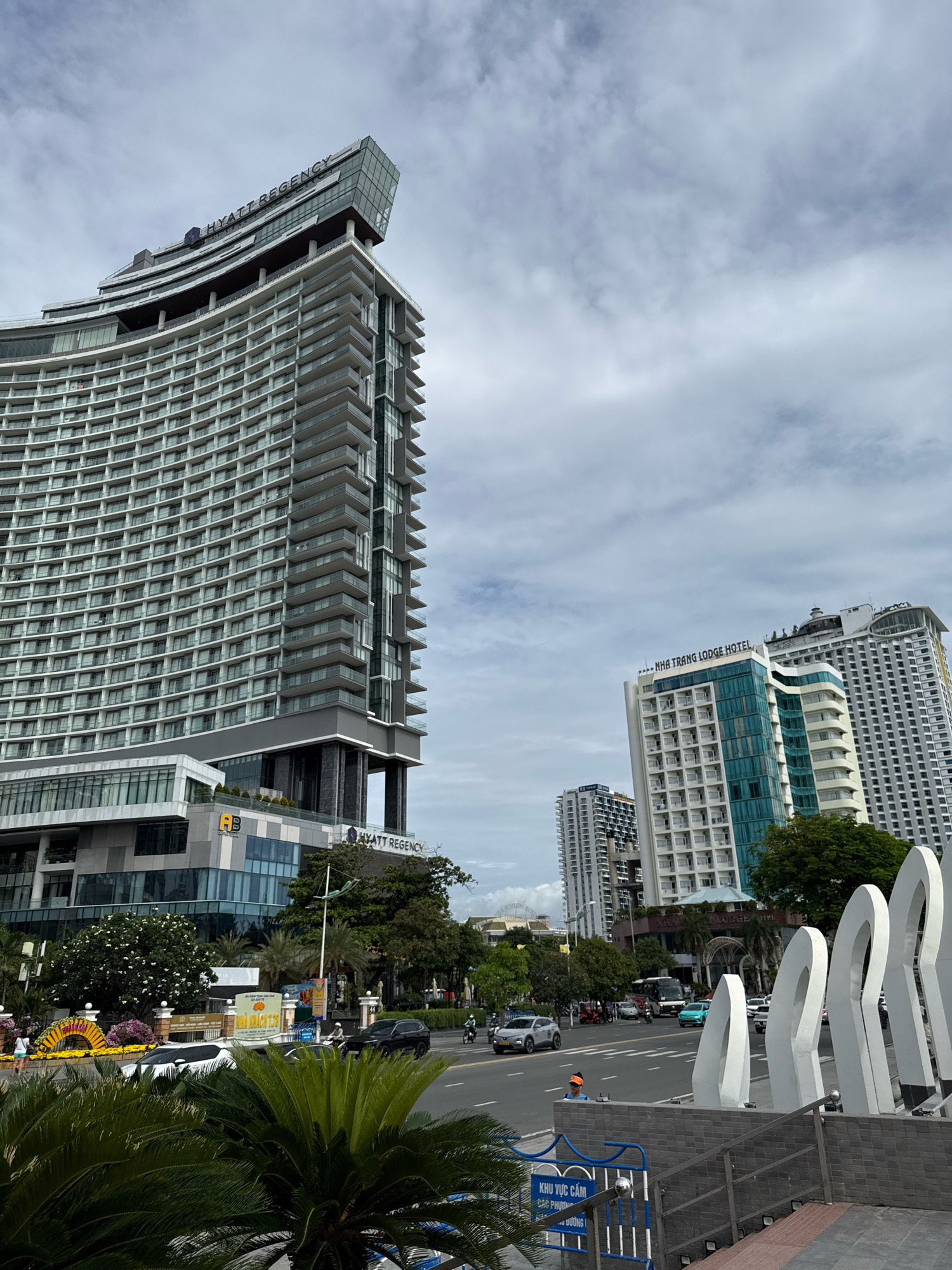 The image captures a street view in Nha Trang, Vietnam, featuring the prominent Hyatt Regency hotel, a curved, modern building with many balconies and a distinctive glass-enclosed top section. Adjacent to it, the Nha Trang Lodge Hotel is visible, as well as other high-rise buildings in the background. A street runs across the foreground with several vehicles, including cars and motorcycles, in motion. In the lower right corner, a section of a public space is visible with distinctive white, arch-like sculptures. Palm trees and decorative floral arrangements are present along the roadside, suggesting a tropical urban environment. The sky is overcast with scattered clouds. A blue and white fence with Vietnamese text is visible in the lower portion of the frame.