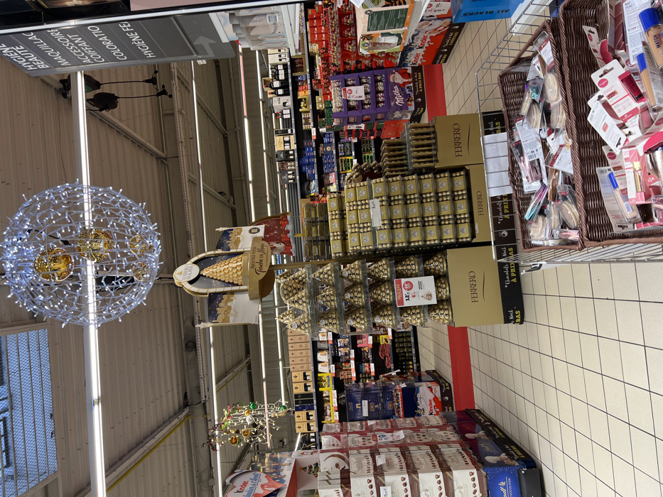 The image displays the interior of a hypermarket or supermarket located in Hérouville-Saint-Clair, France, characterized by holiday-themed displays. A large spherical light fixture composed of numerous white LED lights, containing three larger golden orbs, hangs from the industrial-style ceiling which features exposed utilities and fluorescent lighting strips, as well as a visible skylight. To the left, a smaller decorative wire structure adorned with colorful baubles is present.
The central area is dominated by an extensive Ferrero Rocher chocolate promotion. This includes a tall display stand with a graphic of a chocolate pyramid and the "Ferrero Rocher" brand name, supported by multiple tiered displays of individually wrapped Ferrero Rocher chocolates arranged in pyramid shapes on golden bases. Adjacent to these are large stacks of golden-packaged Ferrero Rocher boxes, with some visible pricing such as "12,99€."
On the left side, stacked cardboard boxes of Lindt chocolates are visible, and further into the background, a "Kinder" branded display is partially seen. The right side of the frame features an overhead sign listing product categories in French: "HYGIÈNE BEAUTÉ" (Hygiene Beauty), "MAQUILLAGE" (Makeup), "ACCESSOIRE COIFFANT" (Hair Accessory), "COLORATIO" (Hair Dye), and "HYGIÈNE FE" (Female Hygiene). Below and behind this sign, shelves are stocked with various beauty and personal care products. Further back, shelves containing bottles of wine and other general merchandise are visible. In the immediate foreground on the right, a wire shopping basket is filled with various packaged cosmetics, including compacts and lip products.
The floor throughout the visible area is a light-colored tiled surface with a grid pattern. A red carpet strip runs down one of the main aisles in the midground. No individuals are discernible within the frame.