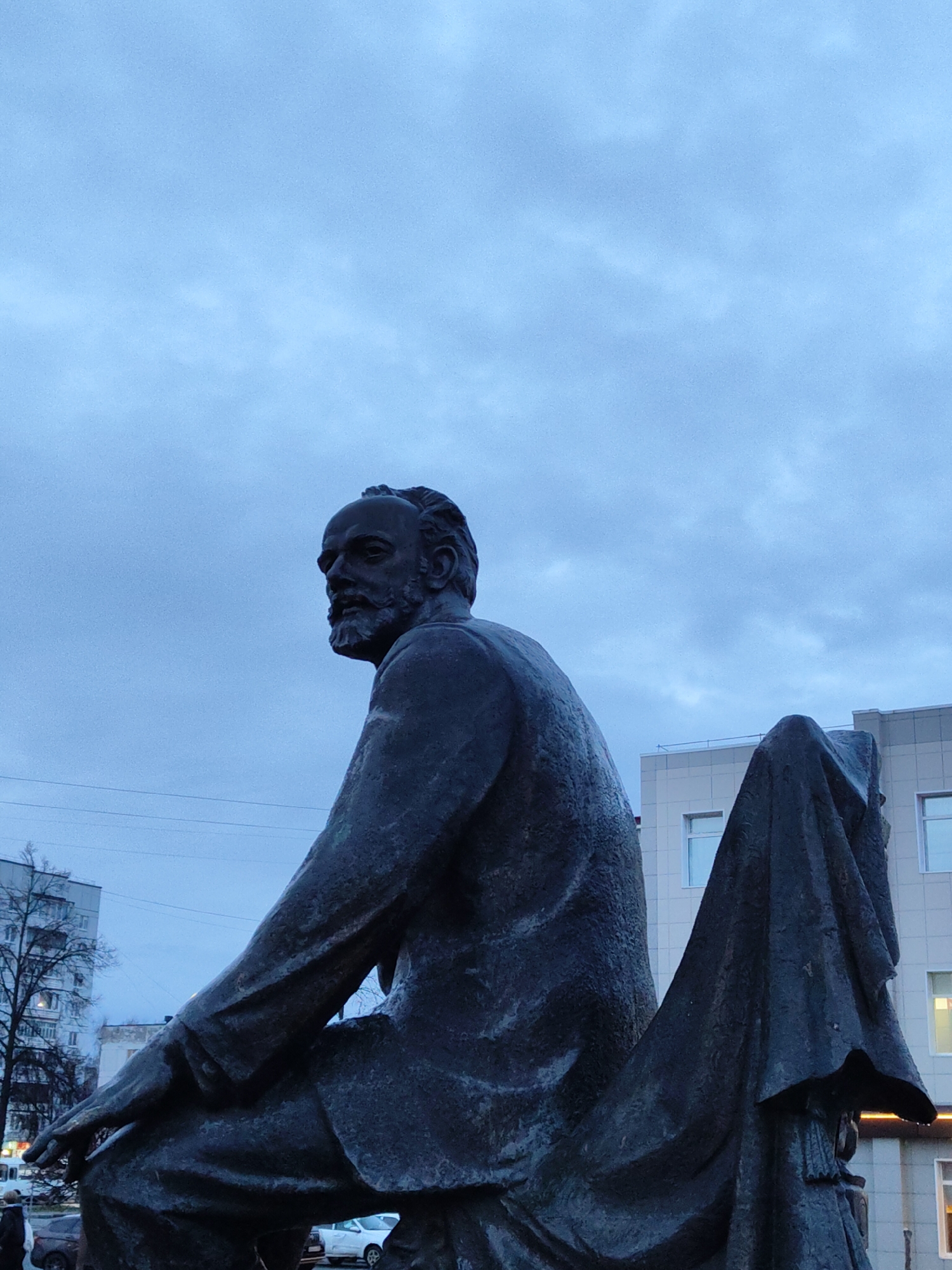 The image captures a seated bronze statue of a man with a beard, facing left, against a backdrop of a city street in Unknown. The man is wearing a suit-like garment. He is seated with his left hand resting on his left knee. Behind the statue is a light-colored building with multiple windows and horizontal lines, and a bare tree with branches visible in the left side. The sky is overcast, and electric wires are visible above the tree. Cars and pedestrians are present on the street.