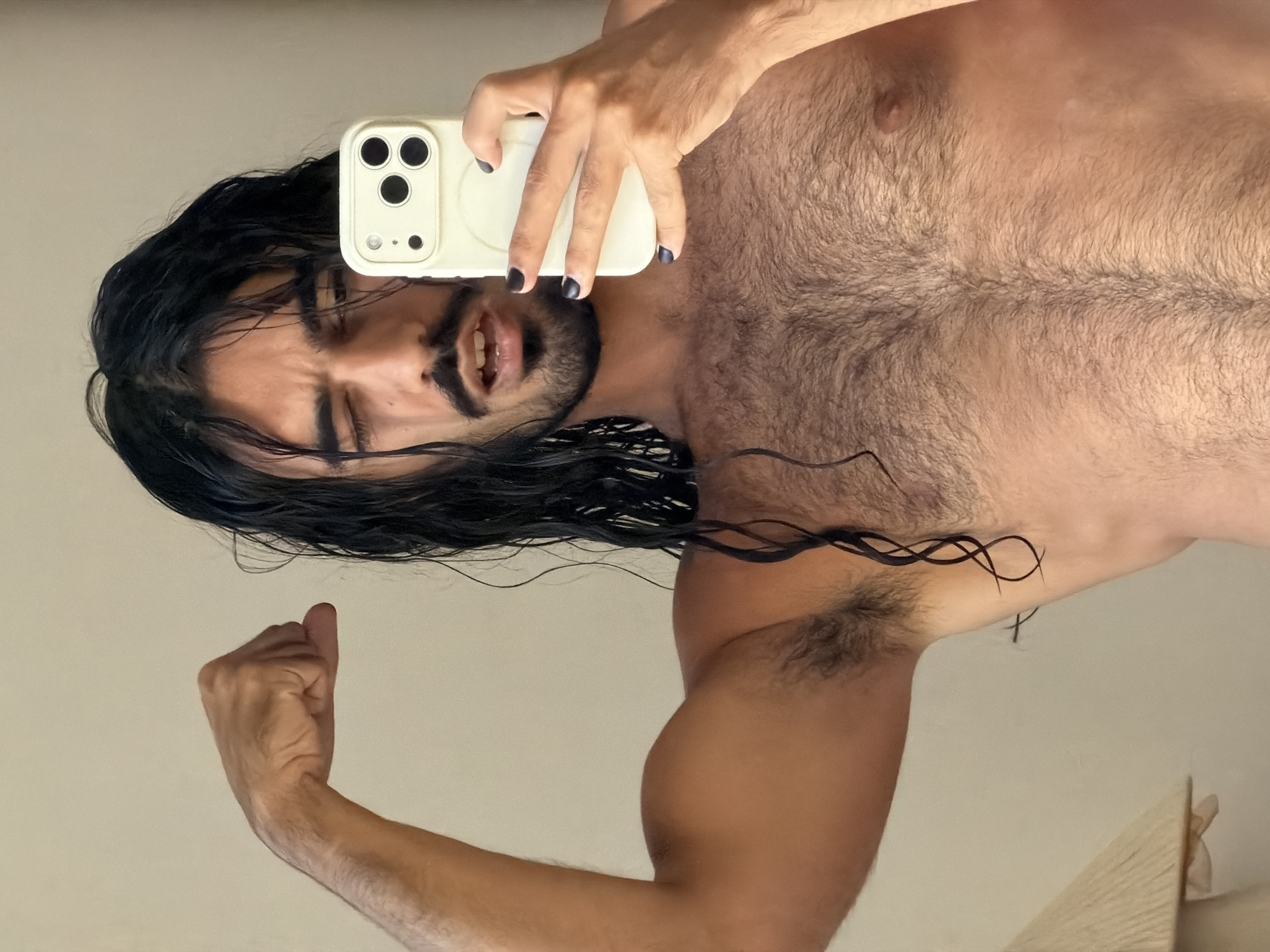 A man with long, dark, wet hair and a mustache is taking a selfie in a room. He is flexing his bicep and winking at the camera. He is holding a white smartphone in his right hand. He has dark nail polish on his fingernails. The man is shirtless and has a hairy chest and armpits. The background is a plain, light-colored wall.
