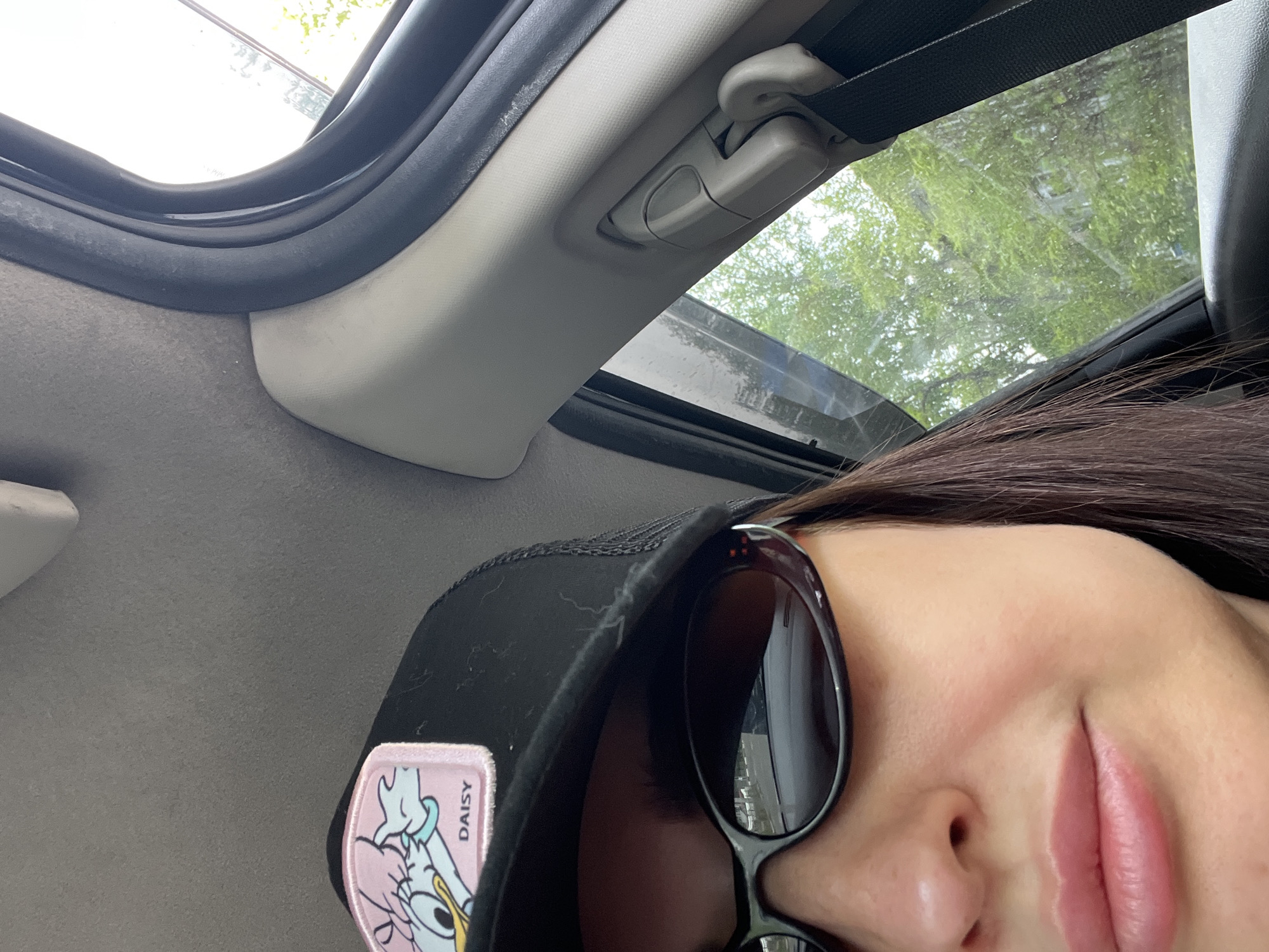 The photograph depicts a partial view of a person wearing sunglasses and a black cap with a Daisy Duck patch inside a vehicle. The vehicle has a sunroof partially open, revealing green foliage outside. The interior of the vehicle is visible, including the roof lining, sunroof frame, and a seatbelt fixture. The location is unknown.