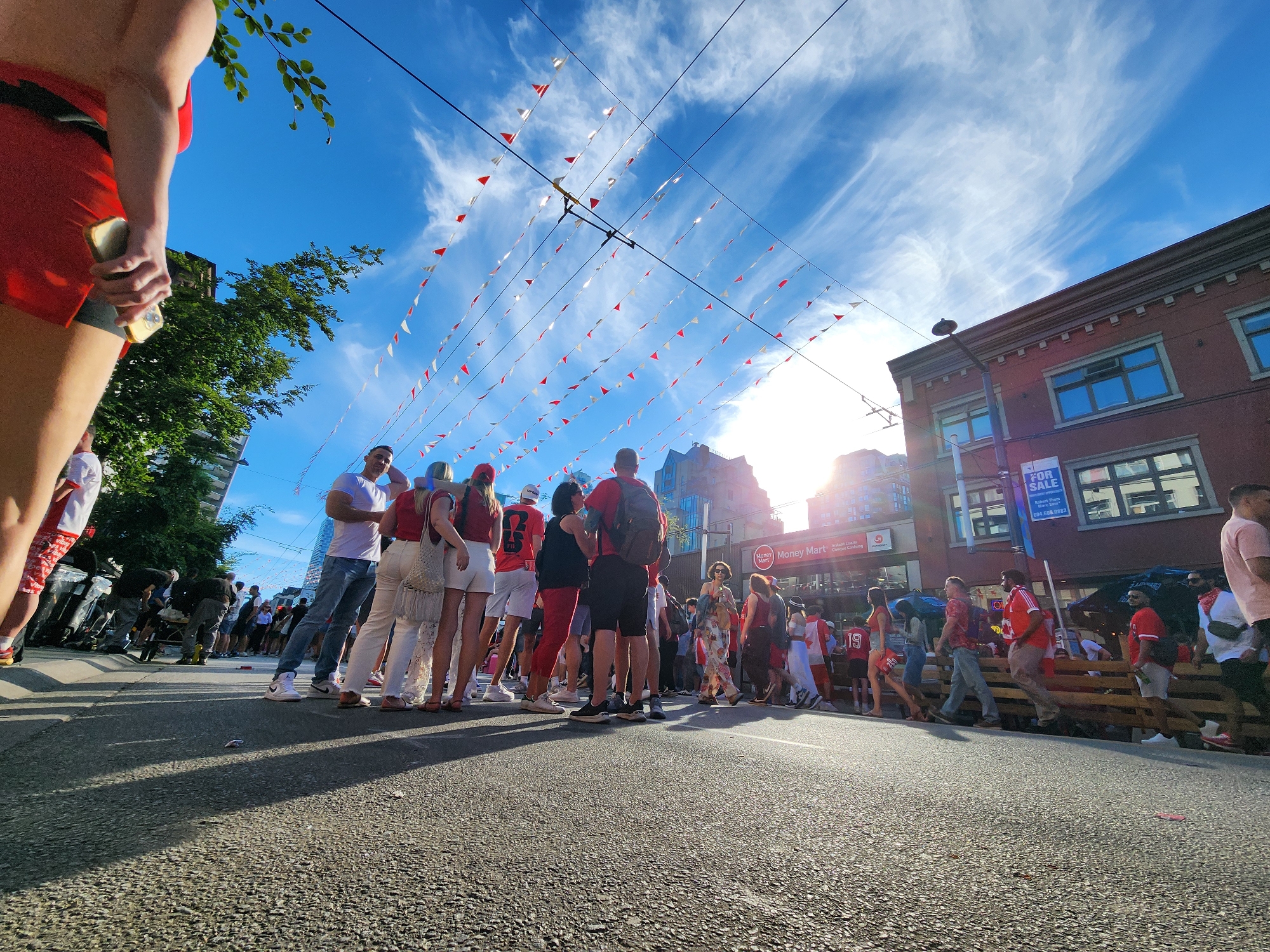 A street scene at eye level, looking up at a crowd of people gathered on a sunny day. The sky is a vibrant blue with wispy white clouds. Red and white triangular flags are strung across the sky, suspended from overhead wires. The crowd is dense, with many individuals wearing red and white clothing, suggesting a sporting event or national celebration. People are walking, standing, and interacting with each other. In the background, buildings line the street, including a brick building with windows and signage that reads "Money Mart." The ground is asphalt, with shadows cast by the people and buildings. A person in the foreground is partially visible on the left, wearing red shorts and holding a smartphone. The overall impression is a lively outdoor gathering on a clear, bright day.