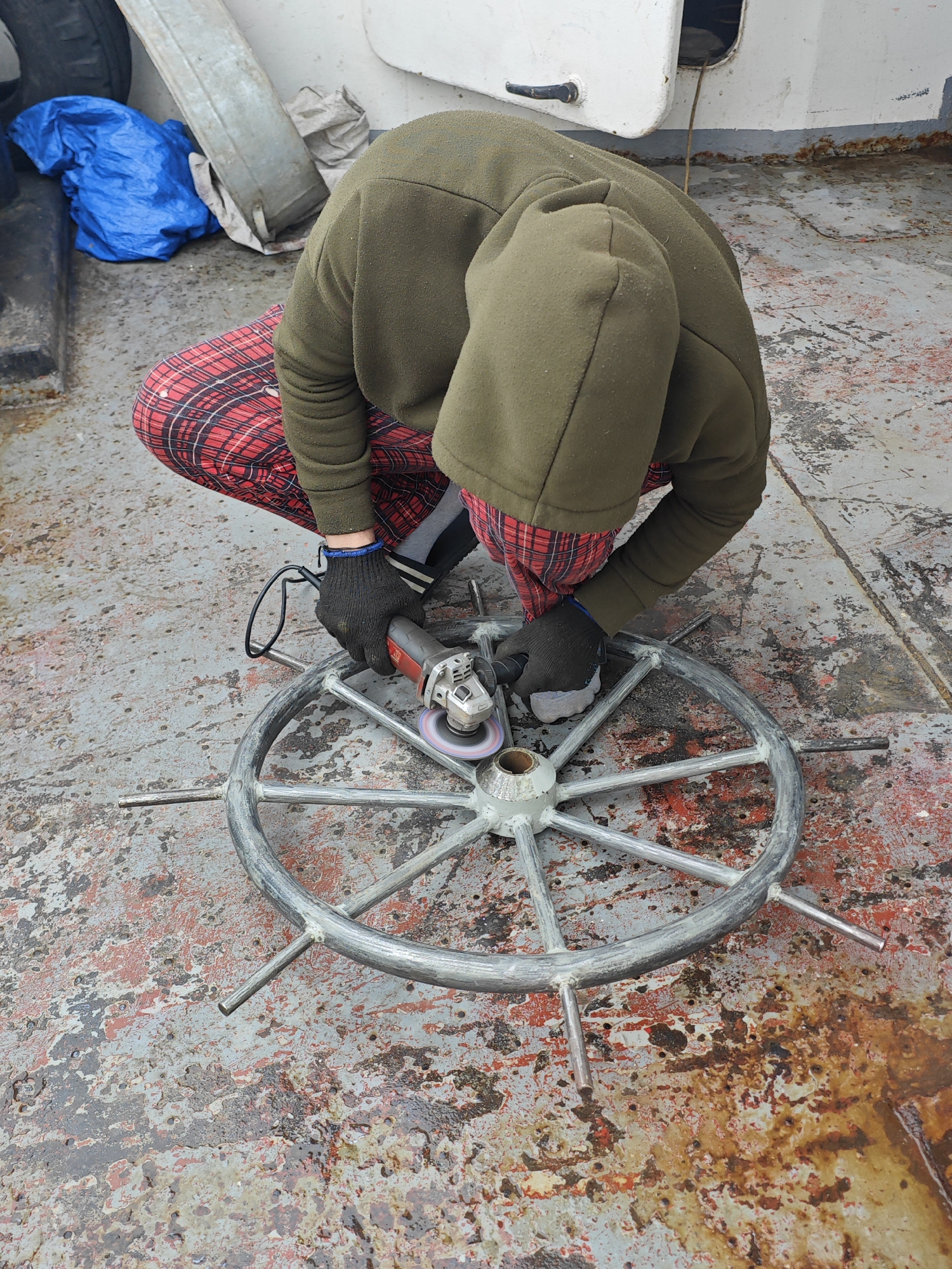 A person is crouched on a weathered, metallic deck, wearing black gloves and a green hooded sweatshirt over a red and black plaid shirt. The individual is using an angle grinder, its cord plugged in, to work on a silver, multi-spoked ship's wheel. The deck surface is stained with rust and what appears to be water. In the background, a blue tarp is visible next to a piece of grey metal. The scene suggests an outdoor work environment, possibly on a boat or dock, in a location like Ермолаево, Russia.