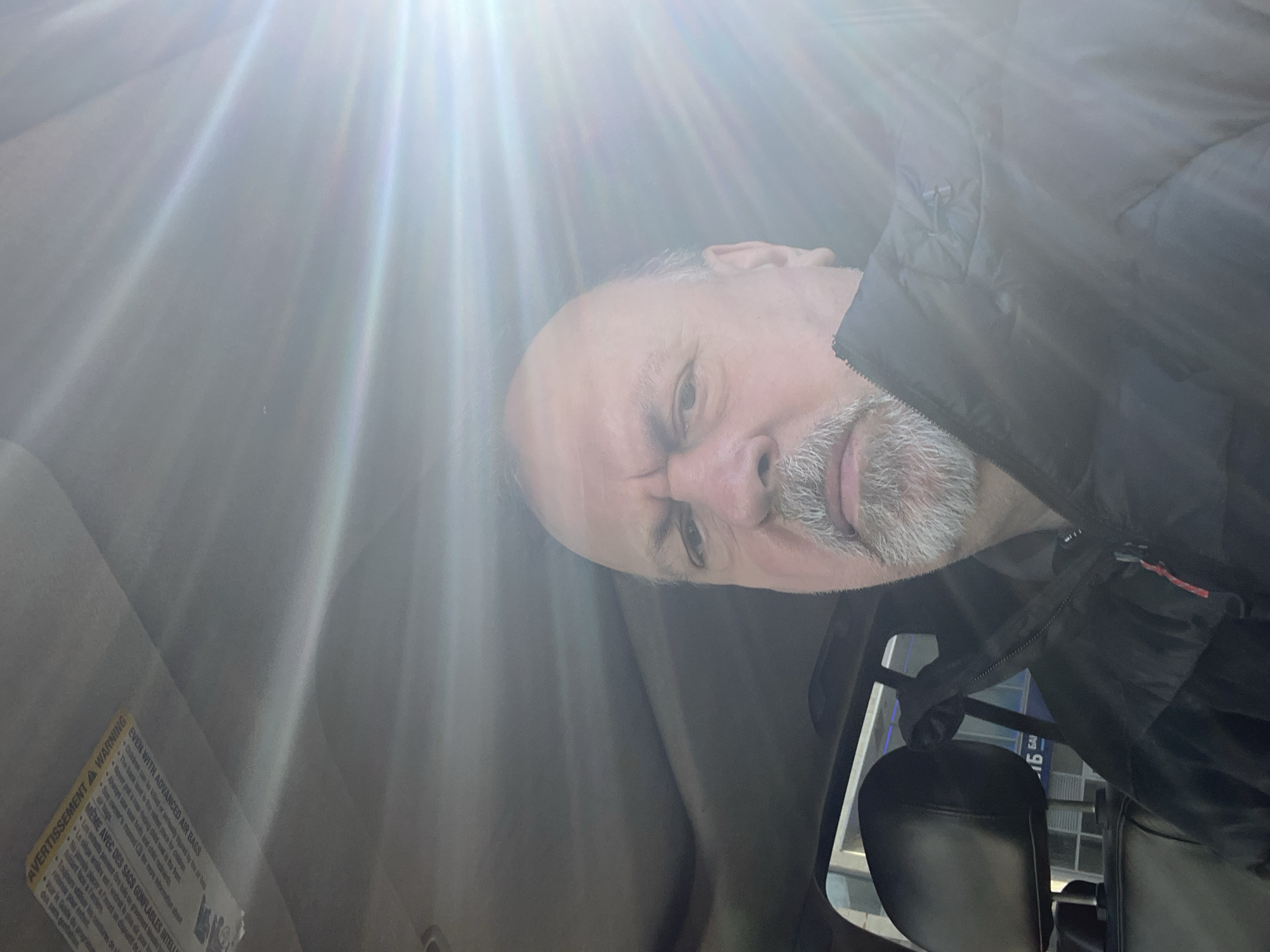 The image appears to be a selfie taken inside a car. The man, who appears to be middle-aged with a bald head and grey beard, is wearing a black jacket. The sun is shining brightly into the car, creating lens flare. A warning label related to advanced air bags is visible on the car's interior. A headrest is partially visible in the background, likely part of the car's seat. There is nothing in the image to indicate the location is Podolsk, Russia.