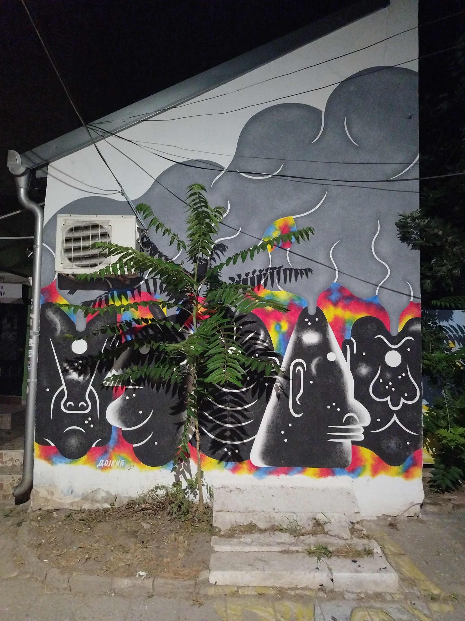 The image shows a building wall covered in graffiti in Novi Sad, Serbia. The graffiti features abstract, monochromatic figures with large heads and simple facial features, set against a background of gray shapes and vibrant, abstract colorful elements. A small green tree grows in front of the wall, partially obscuring the artwork. An outdoor air conditioning unit is mounted on the wall above the graffiti. Wires are visible strung across the upper portion of the wall. The area in front of the wall consists of a dirt patch with some vegetation and a concrete pathway with steps. The lighting suggests it is nighttime or dusk. The graffiti appears to be a mural art piece.