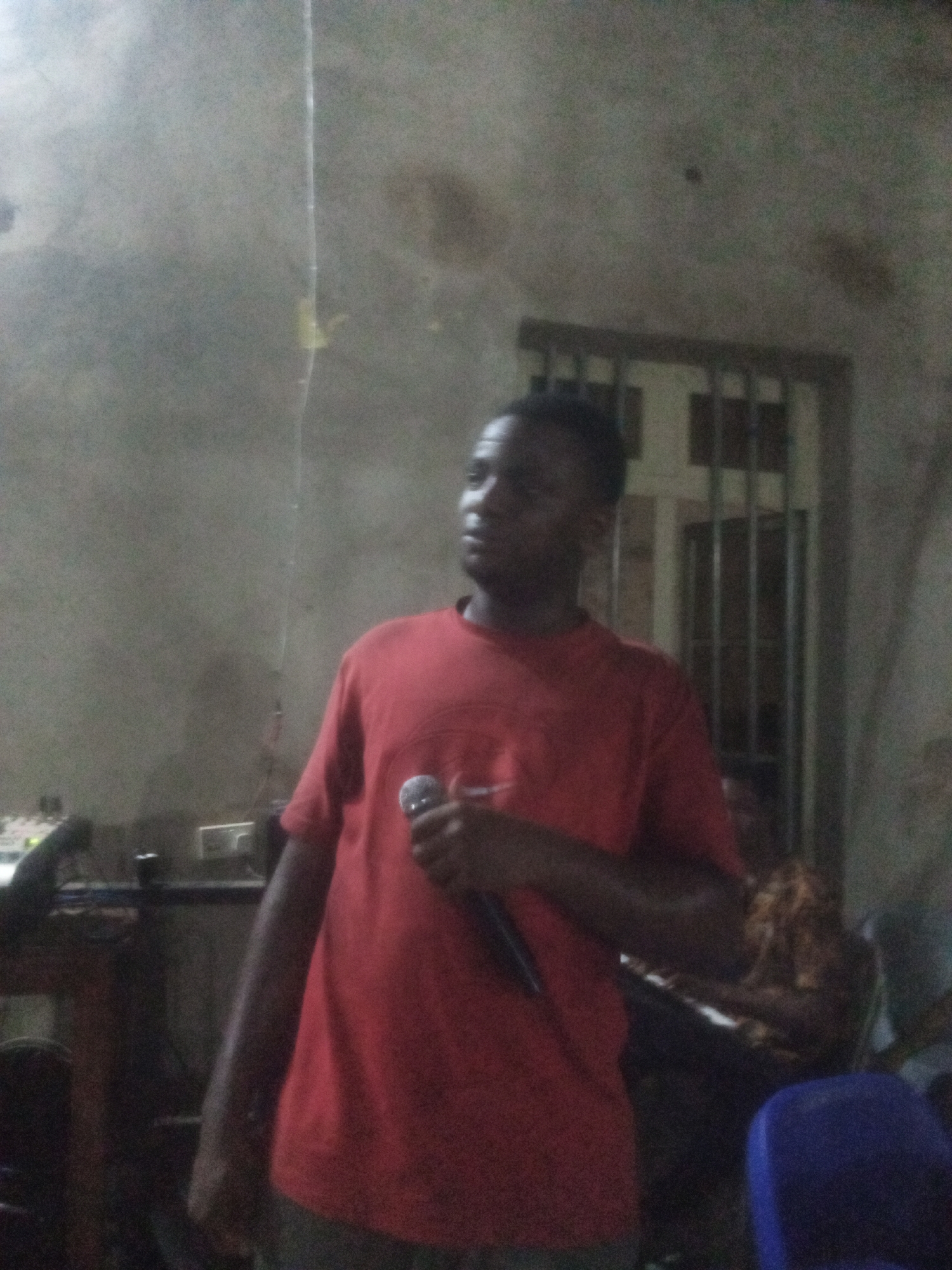 An indoor shot shows a man holding a microphone, wearing a red shirt. The background includes a window with bars, a person seated, and musical equipment. The location is Ukwu-Abwa, Nigeria.