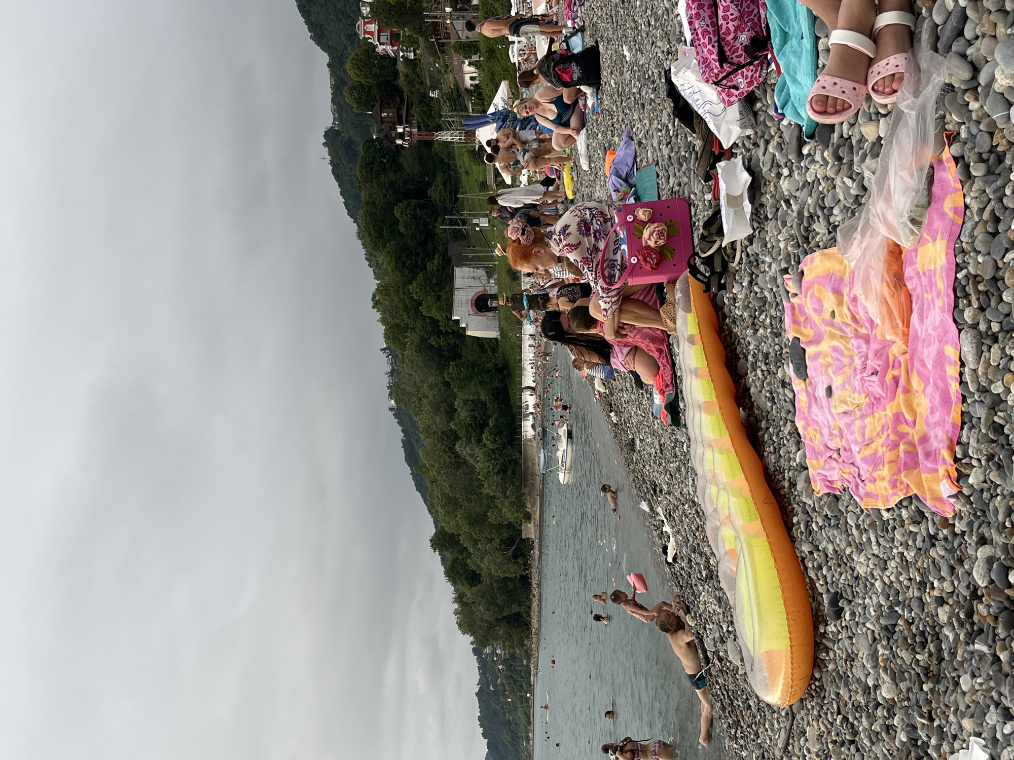The image shows a crowded pebble beach in Sochi, Russia, under an overcast sky. In the foreground, several people are relaxing on the beach. An inflatable raft lies on the pebbles, alongside a folded pink and orange towel and other beach belongings. Further back, a group of people are sitting on the beach, some in swimwear. A few individuals are in the water, which appears to be calm. In the mid-ground, a small white boat is moored in the sea. The background features a hillside covered in dense green trees, with scattered buildings and structures visible on the slopes, including a distinct tunnel entrance on the left and a tower structure on the right. The overall scene depicts a typical summer day at a popular beach resort.