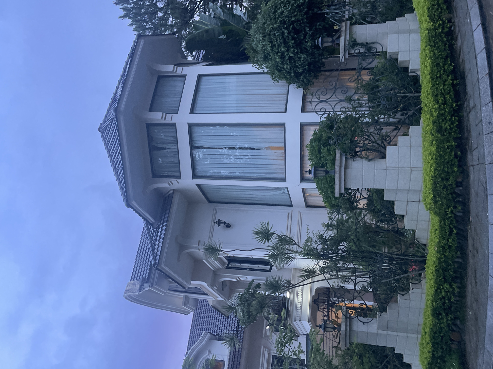 The image depicts a large, modern villa with white walls and a dark tiled roof, set against a twilight sky with subtle purple and blue hues. The villa features prominent bay windows with sheer curtains and decorative architectural details. A manicured garden with lush green hedges and ornamental trees surrounds the property, with a decorative metal fence and stone pillars at the entrance. The scene suggests a residential area in Da Lat, Vietnam, known for its European-inspired architecture and verdant landscapes. No people or specific activities are visible.
