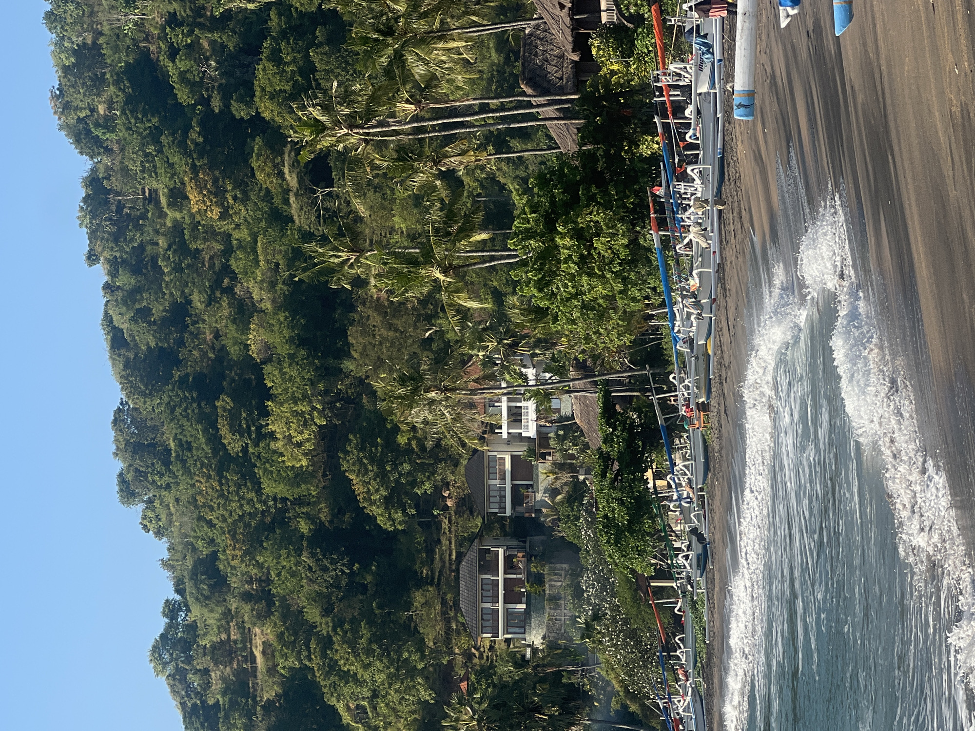 This image depicts a coastal scene in Abang, Indonesia, likely on a beach with dark sand. The foreground features gentle waves rolling onto the shore, with foamy white crests. Along the shoreline, numerous traditional Indonesian fishing boats, known as jukung, are lined up. These boats are predominantly white with colorful outriggers and some have red, blue, or orange poles extending from them.
Behind the boats, a line of lush green trees and palm trees marks the transition to a hillside covered in dense vegetation. Several buildings are visible nestled into the hillside. Two prominent multi-story structures with balconies and dark tiled roofs stand out, appearing to be villas or residences. Further back, more trees and vegetation cover the elevated terrain. The sky is a clear, bright blue, suggesting a sunny day. The overall atmosphere is serene and tropical, characteristic of a coastal village in Indonesia.