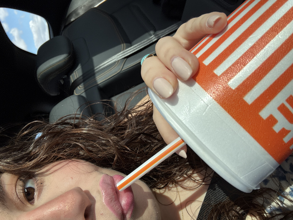 A woman with brown curly hair sips from an orange-striped Whataburger cup using a straw. She is inside a car; black leather seats are visible in the background, along with a glimpse of a blue sky. Her fingernails are painted a neutral color. A turquoise ring adorns her finger. The image focuses on her face and the cup.