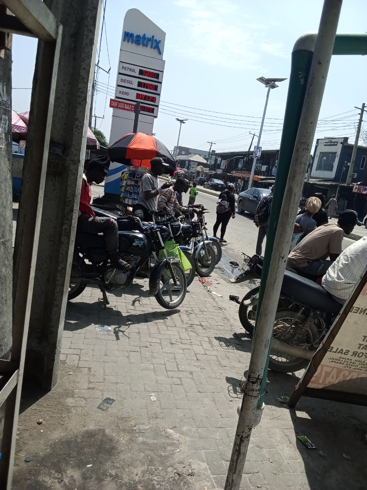 The image depicts a street scene in Ado, Nigeria, likely near a gas station. In the foreground, several motorcycles are parked on a paved walkway. A man wearing a red long-sleeved shirt and dark pants is seated on one of the motorcycles, facing left. To his right, another man in a grey shirt and dark pants is standing. Further down the row of motorcycles, more people are visible, some standing and others seated. Behind the motorcycles, a large sign for "Matrix" gas station displays prices for petrol, diesel, and kero. An orange and black umbrella is open near the gas station sign. The street itself is lined with buildings, some of which appear to be shops. Several cars are visible on the road in the background, and there are power lines overhead. The overall impression is a busy urban environment on a sunny day.