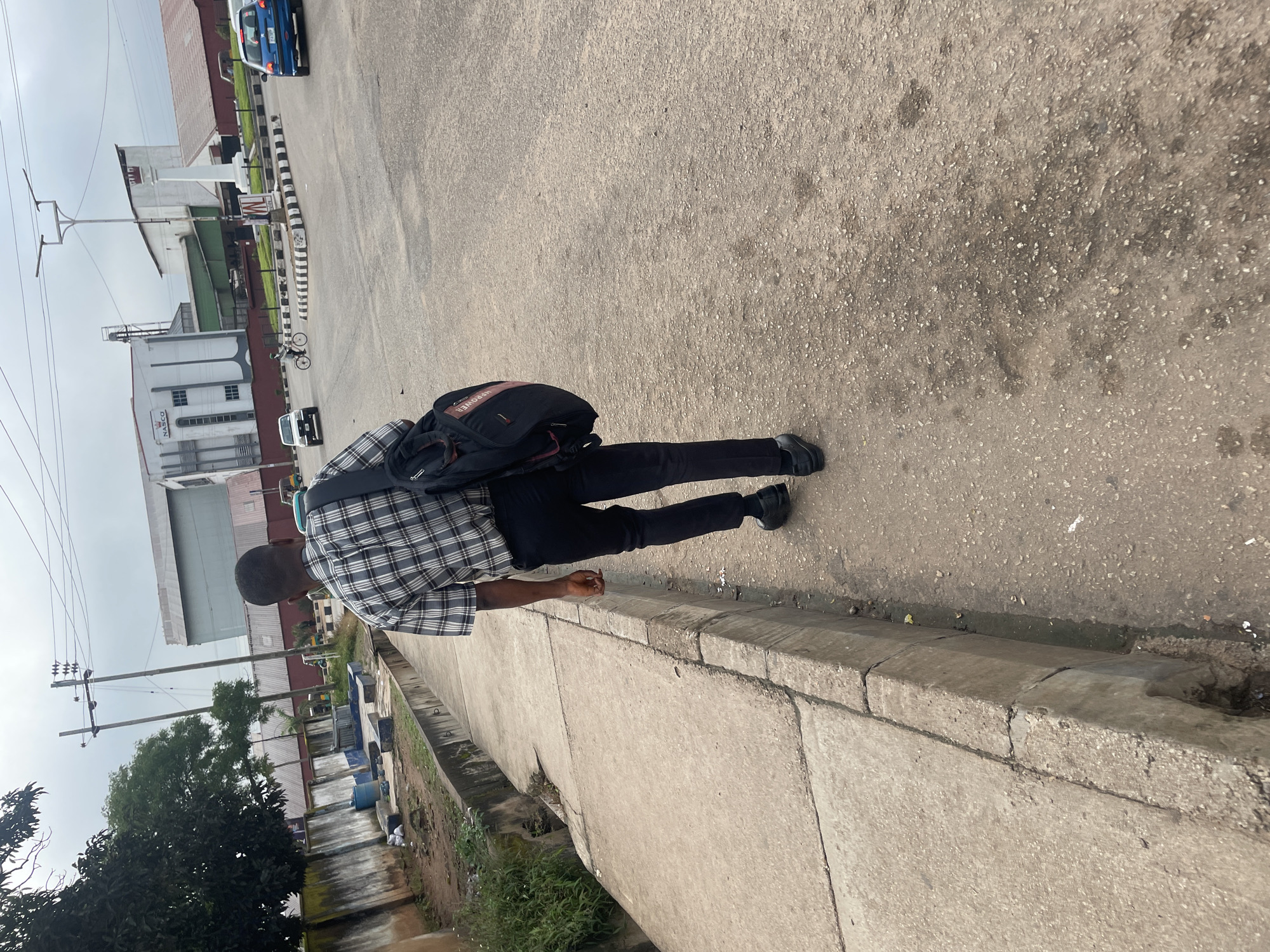 A person is walking away from the camera on a concrete sidewalk next to a street. The person is wearing a black and white plaid shirt, black pants, and black shoes, and is carrying a black backpack with red accents over their right shoulder. The sidewalk is cracked and worn, with a drainage ditch running alongside it. In the background, there are buildings, utility poles, power lines, and several vehicles on the street, including a blue car, a white van, and a tricycle. The sky is overcast. The scene appears to be in an industrial or commercial area.