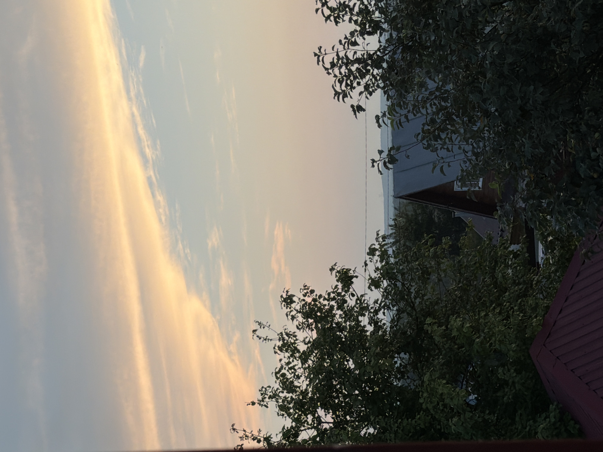 The image depicts a sunset scene over what appears to be a rural or suburban area. The sky is filled with soft, pastel-colored clouds, primarily in shades of peach and pale blue, suggesting the golden hour. In the foreground, lush green trees with dense foliage frame the view. Partially visible structures, including a dark red, pitched roof of a building and the dark grey, angled roof of a house with a visible window, are nestled among the trees. The overall impression is one of tranquility and the end of a day in a natural or semi-natural setting. The location is identified as Nizhnekamsk, Russia.
