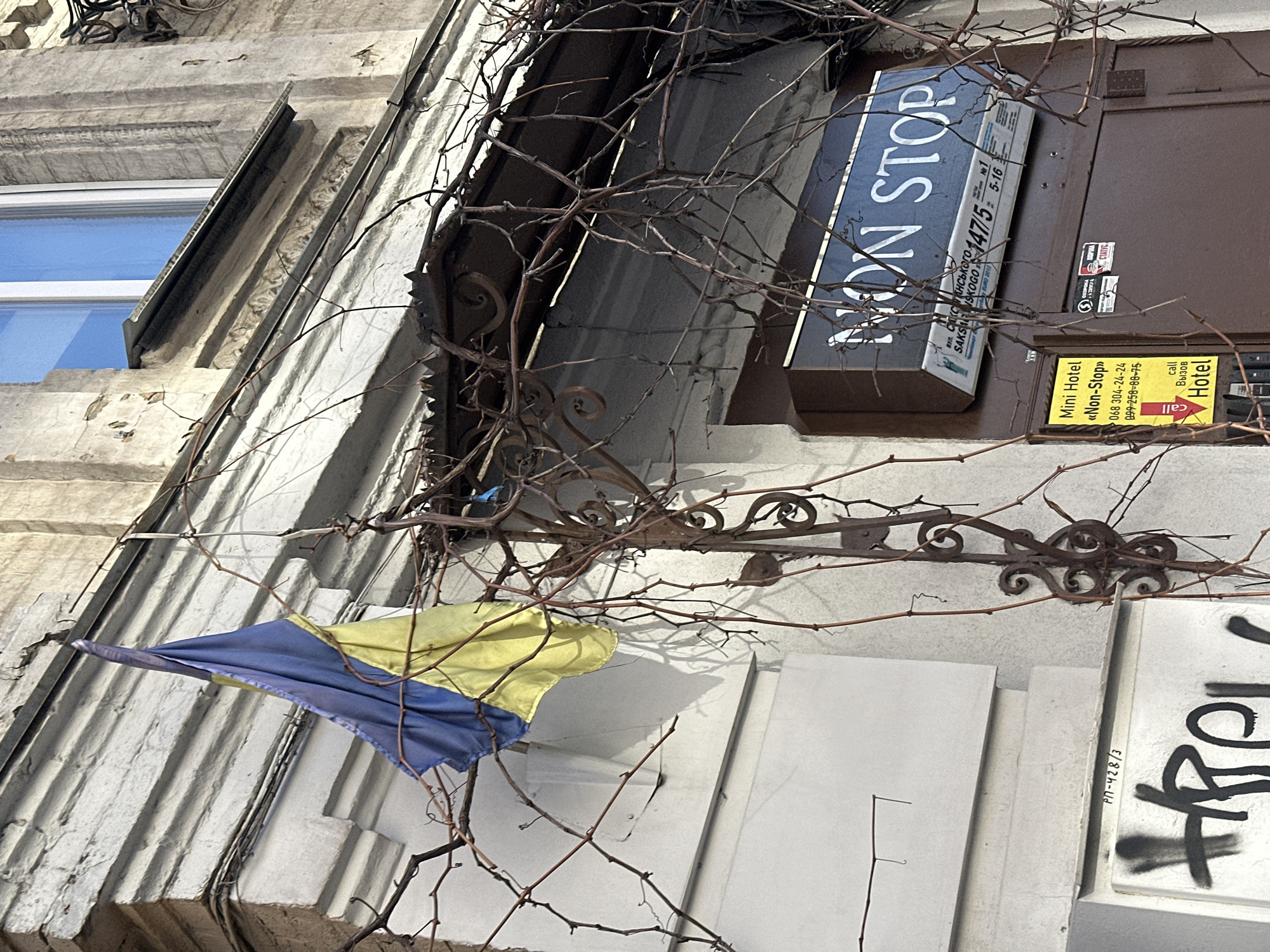 The image depicts the exterior of a building in Kyiv, Ukraine. A weathered Ukrainian flag is prominently displayed. The building has a sign for "Non Stop" mini-hotel at 147/5 Saksahans'koho Street. The building's facade shows signs of wear and tear, with some graffiti. Dormant vines cover portions of the structure, including the sign and an ornamental wrought-iron feature. The scene has a subdued, somewhat neglected aesthetic.