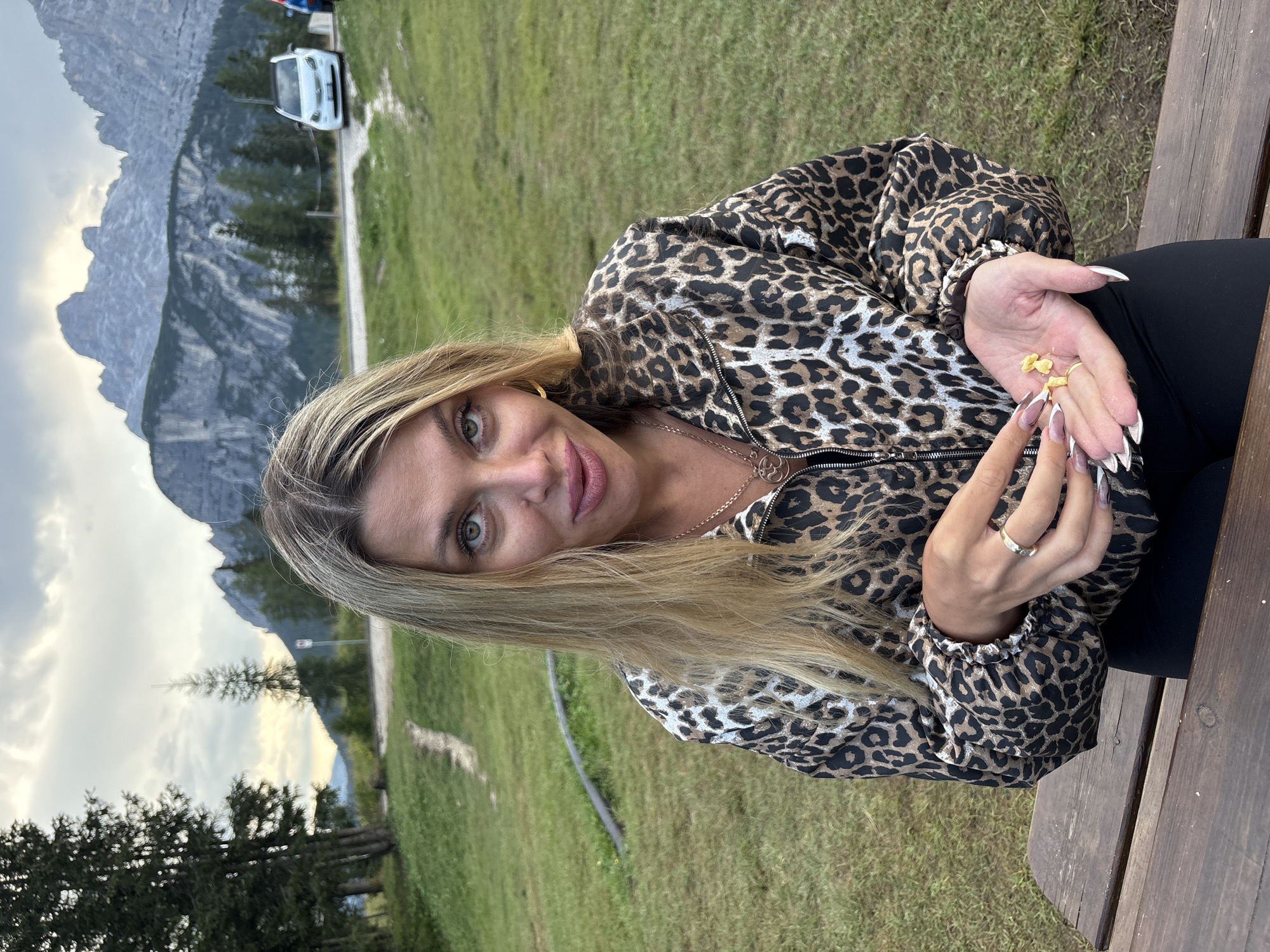 A woman with long blonde hair sits at a wooden picnic table outdoors, holding small yellow objects in her open palm. She is wearing a leopard-print jacket over black clothing and jewelry including rings and a necklace. Her fingernails are long and painted white with dark tips. The background features a grassy slope with a dirt path, trees, a road with a white car, and a large, rugged mountain range under a cloudy sky. The scene appears to be in a natural, mountainous setting, possibly near Auronzo di Cadore, Italy, given the prominent Dolomite mountain peaks.