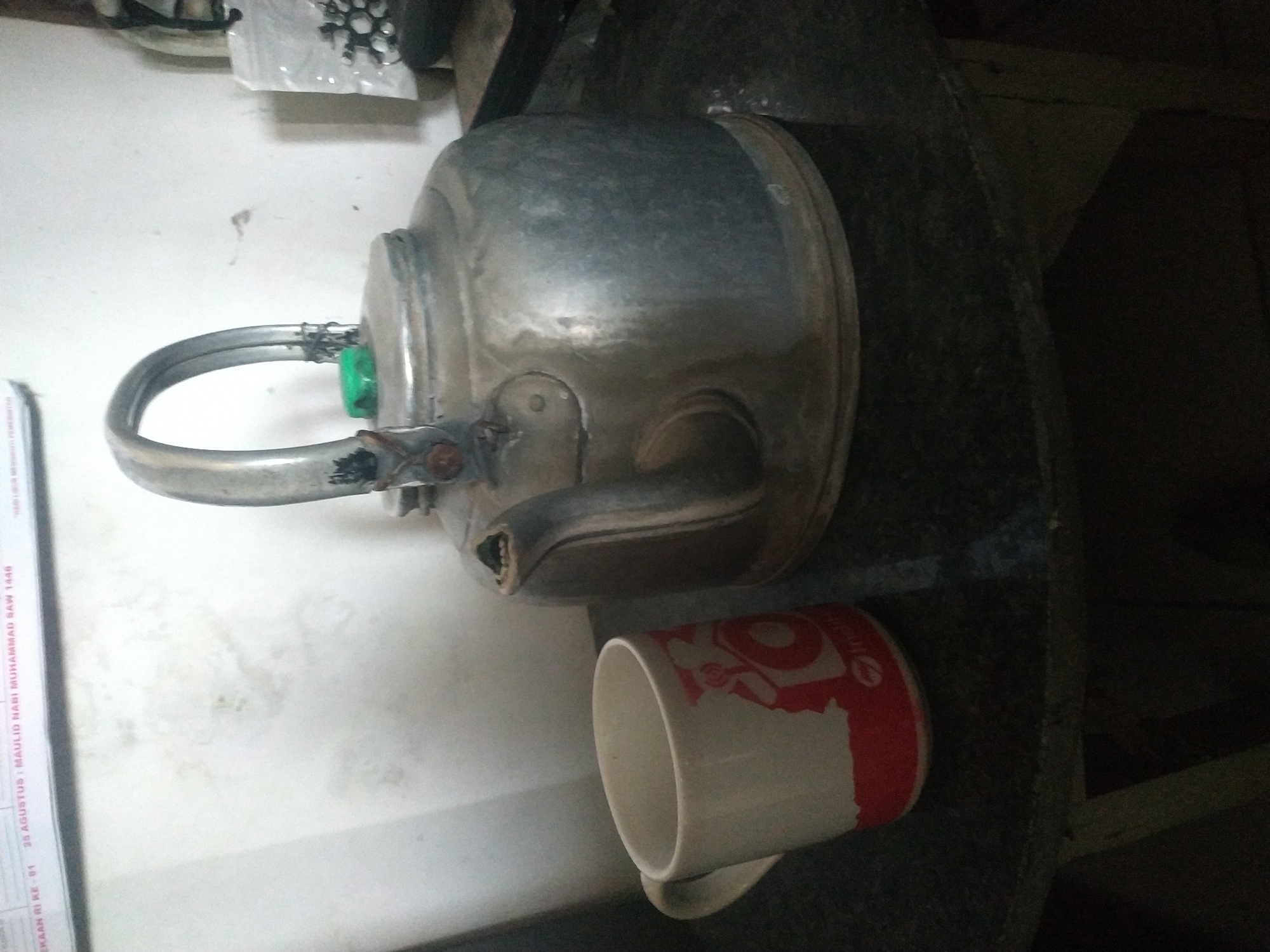 A close-up shot captures a traditional metal kettle and a white mug with red accents, positioned on a dark, reflective surface, possibly a table. The kettle, with its arched handle and spout, appears to be made of aged aluminum or a similar metal, showing signs of wear and tear. A small green cap is visible on top of the kettle. To the left of the kettle sits a white mug with a red design wrapping around its base, featuring a circular logo with text. The background is a plain, light-colored wall, with some faint marks and stains suggesting it's an interior space. A blurred poster is partially visible at the top edge of the frame, displaying text that appears to be in Indonesian, including the date "25 AGUSTUS". The overall lighting is dim, with the subjects casting shadows on the reflective surface. Based on the Indonesian text on the poster, this scene is likely located in Arjasa, Indonesia.