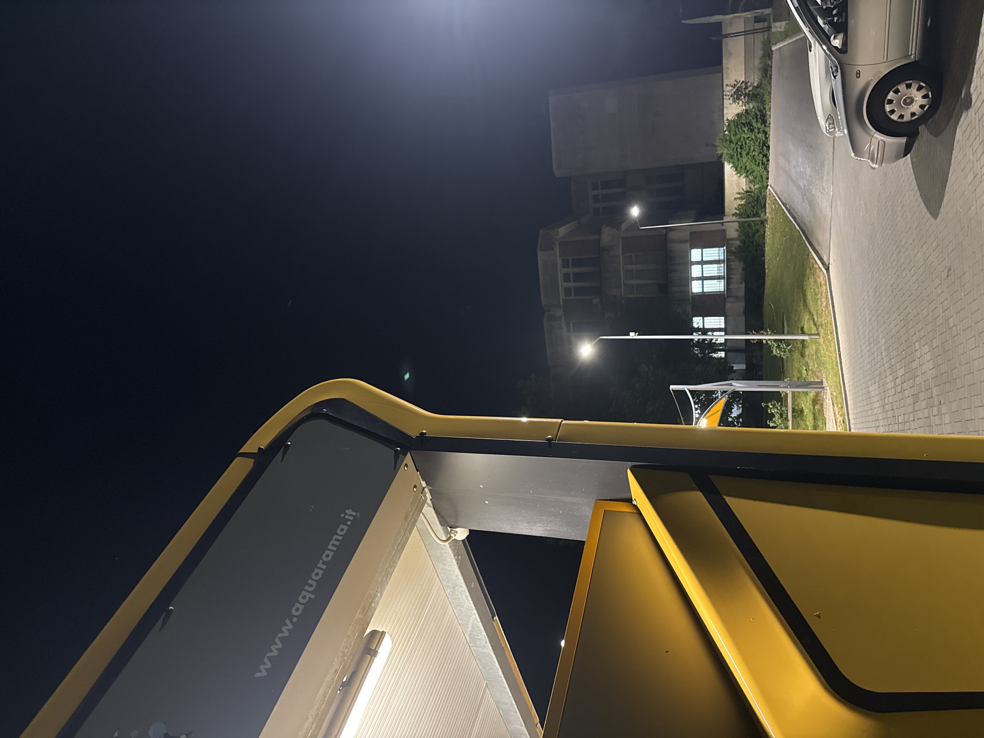 This is a night-time, eye-level, medium shot image taken in Pleven, Bulgaria. In the foreground, a bright yellow structure with black trim dominates the left side of the frame, its reflective surface catching the light. A white linear light fixture is visible on the underside of the structure's canopy. The website "www.aquarama.it" is inscribed on a dark panel above the yellow structure.
To the right, a silver car is parked on a paved driveway. Beyond the car, a building with a concrete facade and multiple windows is illuminated by streetlights. Trees and green grass surround the building, and another streetlight stands to the left of the building. The sky is dark, suggesting it is nighttime. The overall scene appears to be an outdoor area, possibly near a car wash or service station, given the prominent branding and car.